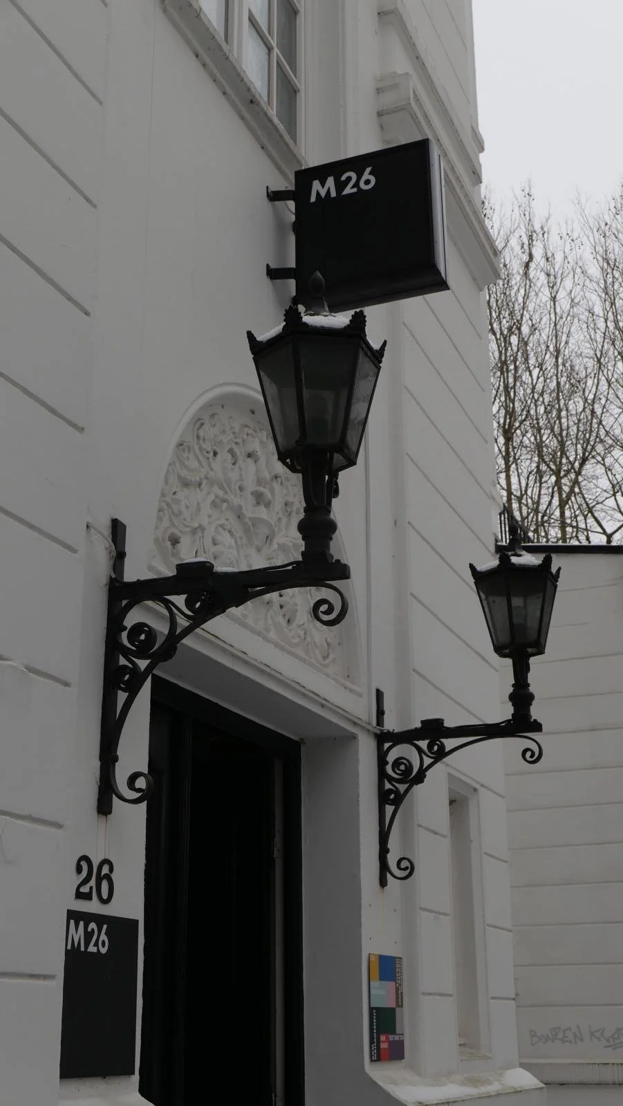 Außenansicht eines weißen Gebäudes mit schwarzem Straßenlaternen- und Ladenschilder, Nummer 26, im Winter mit Schnee.