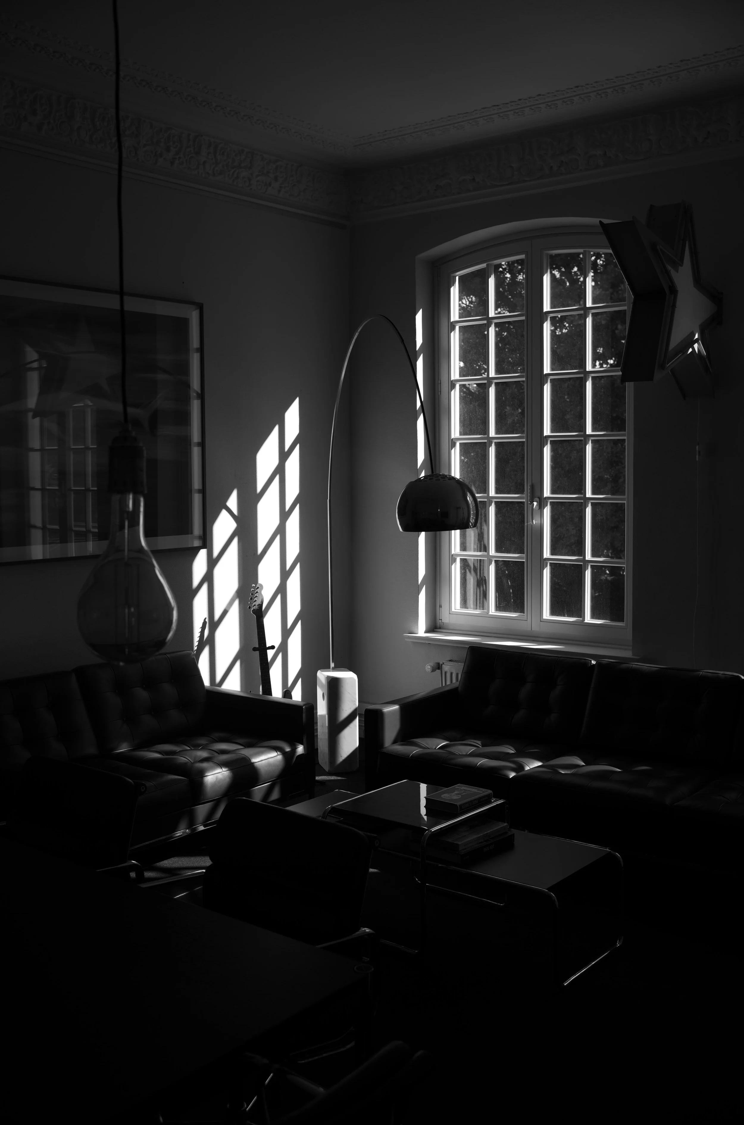 Ein schwarz-weiß Foto eines Wohnzimmers mit Sonnenlicht, das durch ein großes Fenster scheint. Im Raum sind zwei Ledersofas, ein Couchtisch mit Büchern, eine Stehlampe und eine Gitarre im Hintergrund zu sehen. Es gibt auch dekorative Sterne an der Wand.