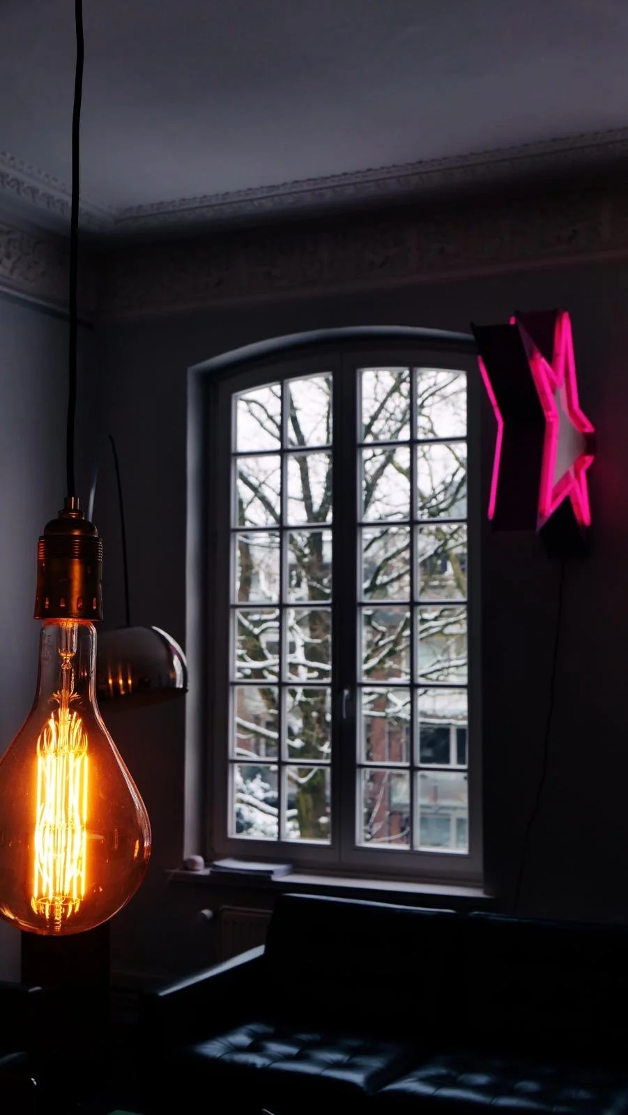 Ein heller, nostalgischer Edison-Glühbirnen-Lampen hängt an der Decke vor einem Fenster, durch das schneebedeckte Bäume und Gebäude sichtbar sind. An der Wand sind eine goldene, metallene Lampe und eine neonrosa-leuchtende Sterne-Form angebracht.