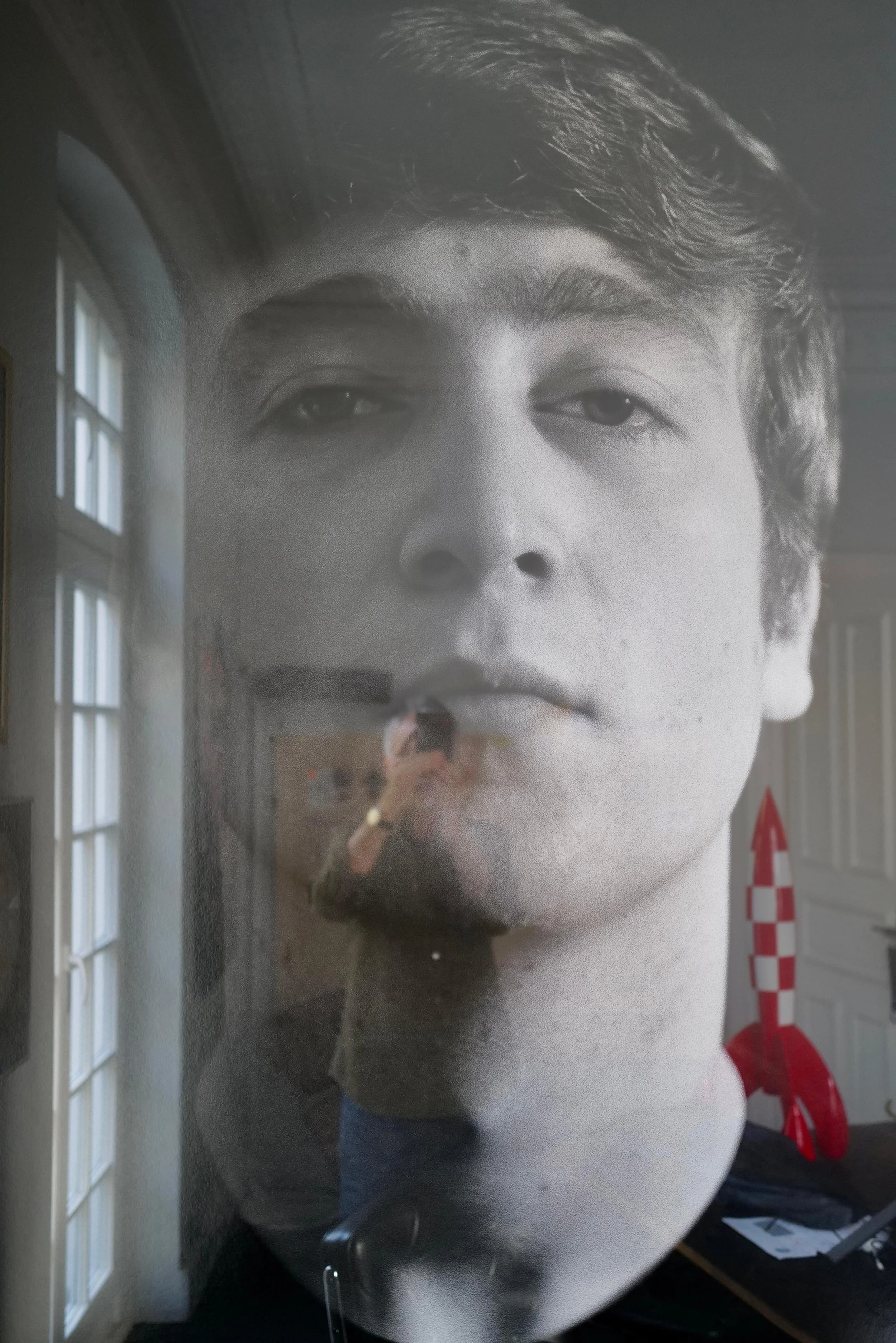 Großes schwarzes und weißes Poster eines jungen Mannes mit schwarzen Haaren, hängt an einer Wand, reflektiert einen Raum mit Fenstern und einer Person, die ein Smartphone hält.