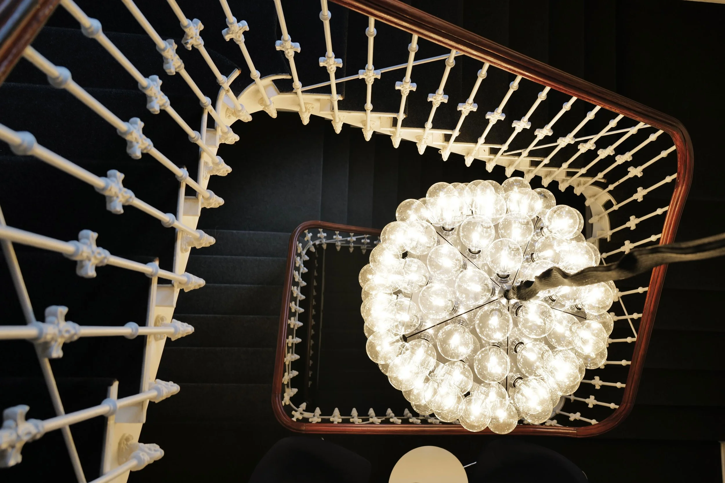 Blick nach unten auf eine moderne Treppe mit weißen Geländerstäben und schwarzen Stufen, im Zentrum hängt eine große Kugel aus vielen Glühbirnen, die hell leuchten. Flos Lampe