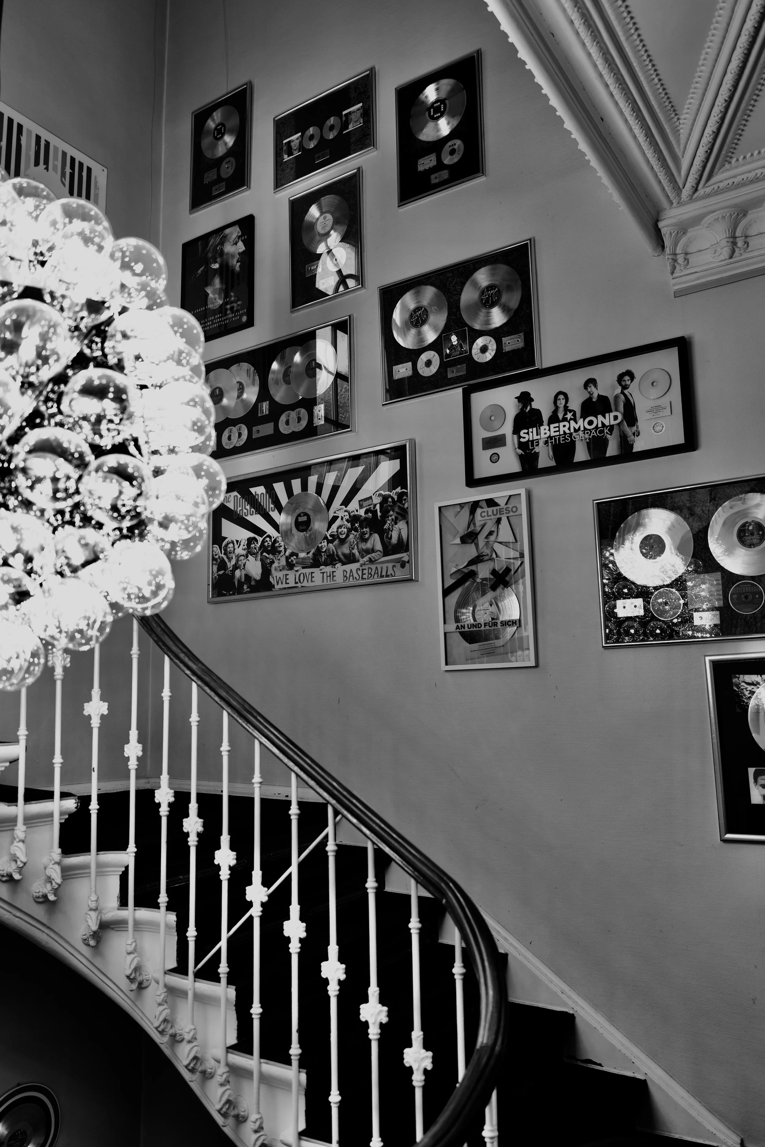 Schwarze und weiße Fotowand mit Musikauszeichnungen und Plakaten, teilweise in Rahmen, in einem eleganten Raum mit Kronleuchter. Perspektive auf eine Treppe mit weißem Geländer.