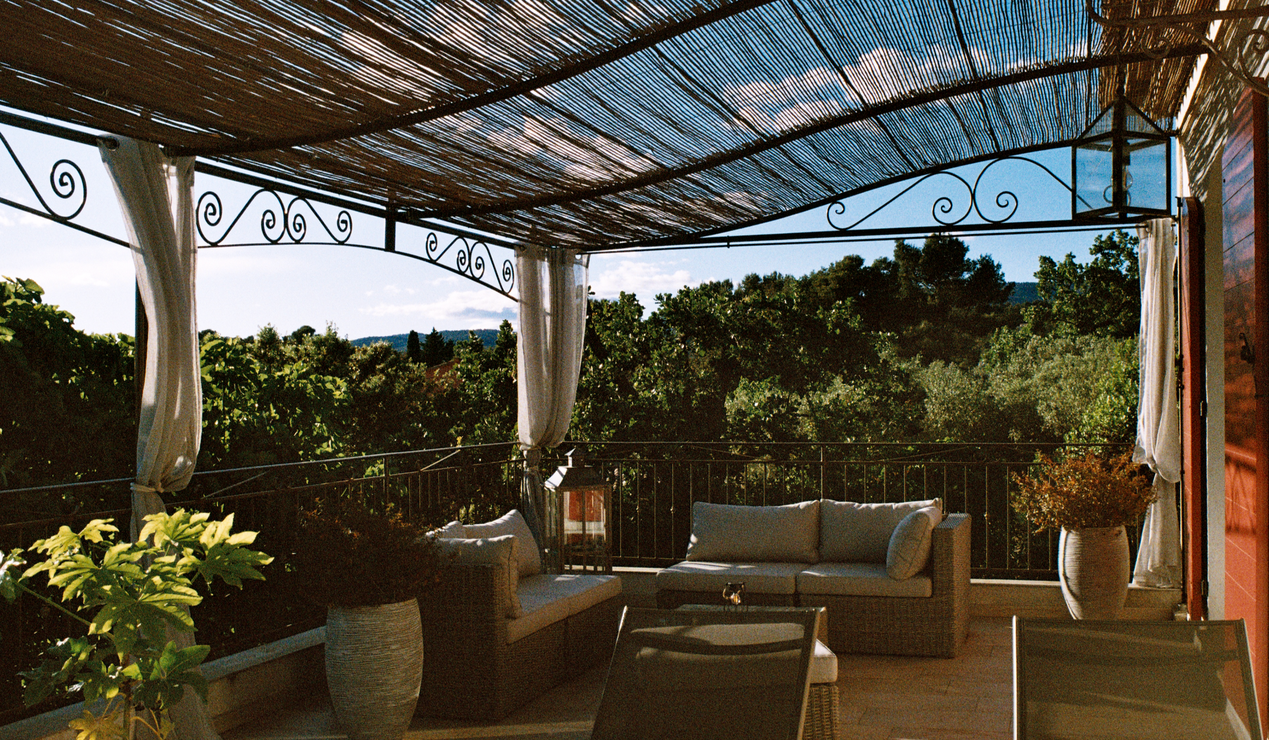 Une terrasse privative avec une pergola en bois, mobilier d'extérieur en rotin avec coussins, plantes vertes, une porte avec volets rouges, vue sur un paysage de campagne avec des arbres et des montagnes au loin.