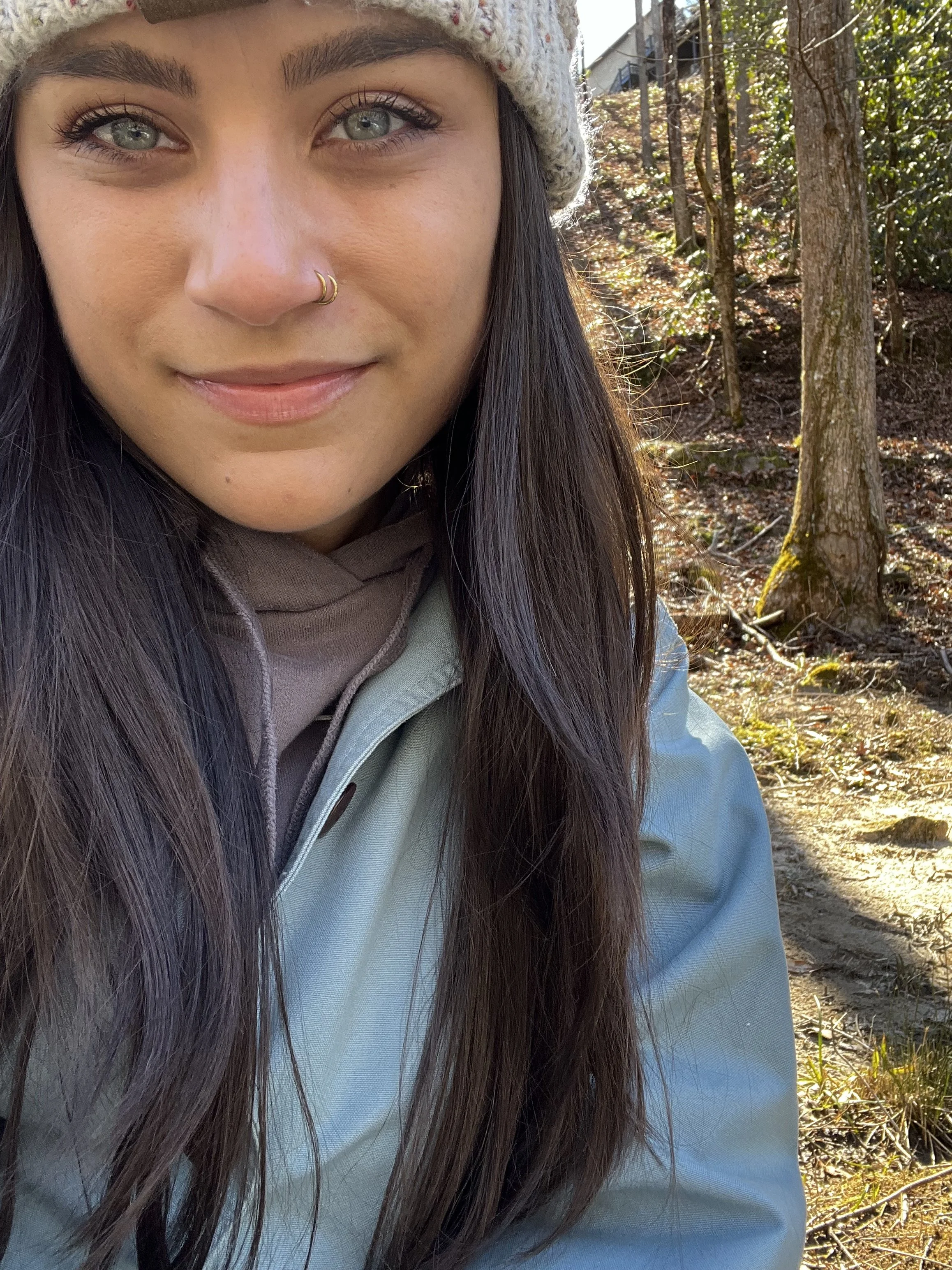 Close-up of a young woman with long dark hair, light blue eyes, wearing a gray beanie, nose ring, and outdoor clothing, smiling in a forest setting with trees and sunlight in the background.