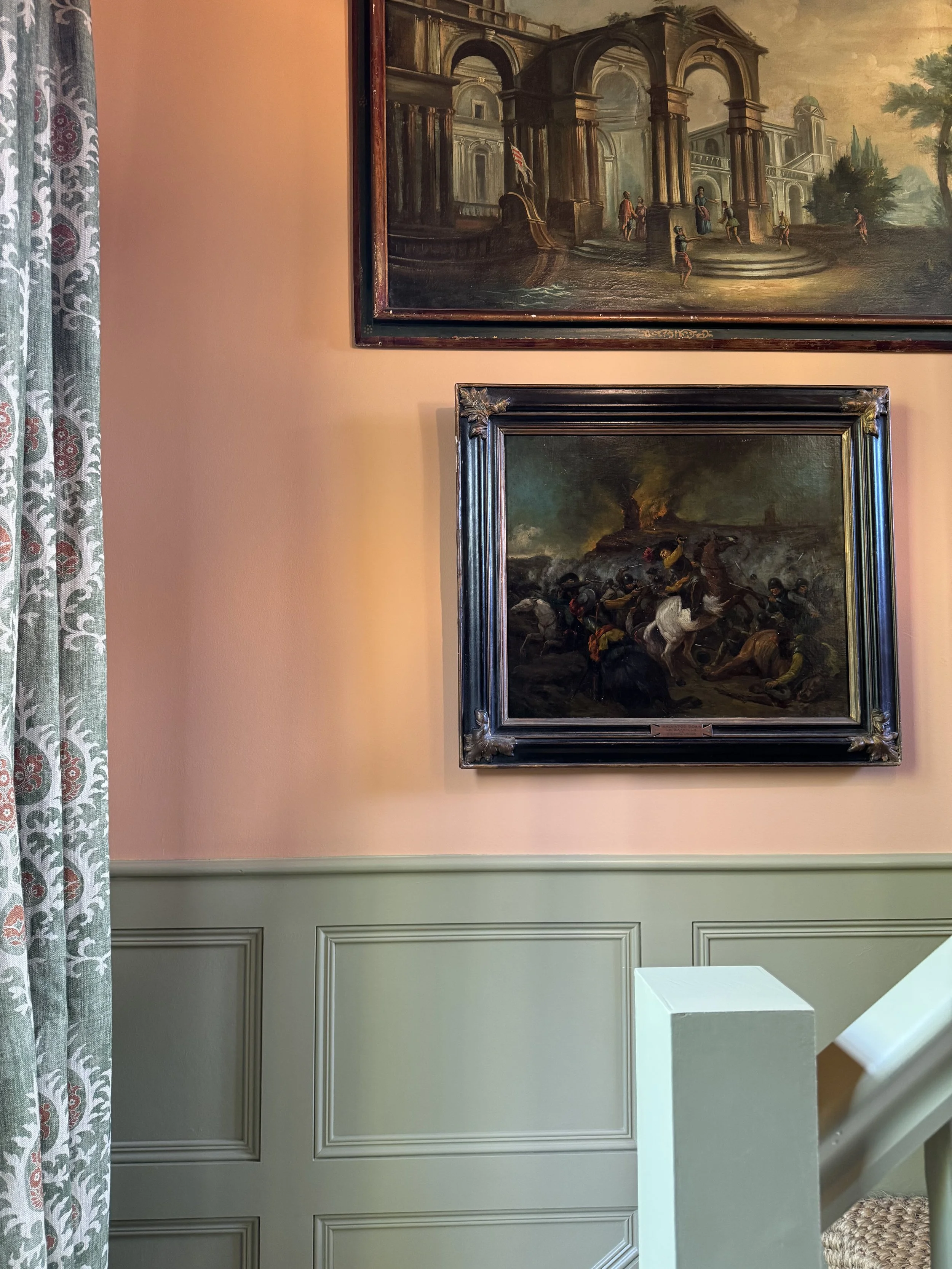 Interior of a room with pink walls, two framed classical paintings, a green wainscot, and patterned curtains.