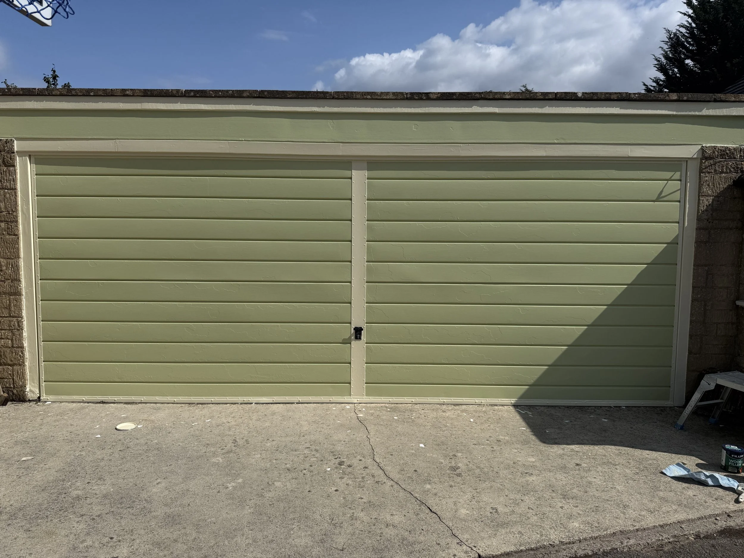 Light green garage door with horizontal panels, surrounded by brick and stone walls, on a paved driveway with some tools and paint cans nearby.