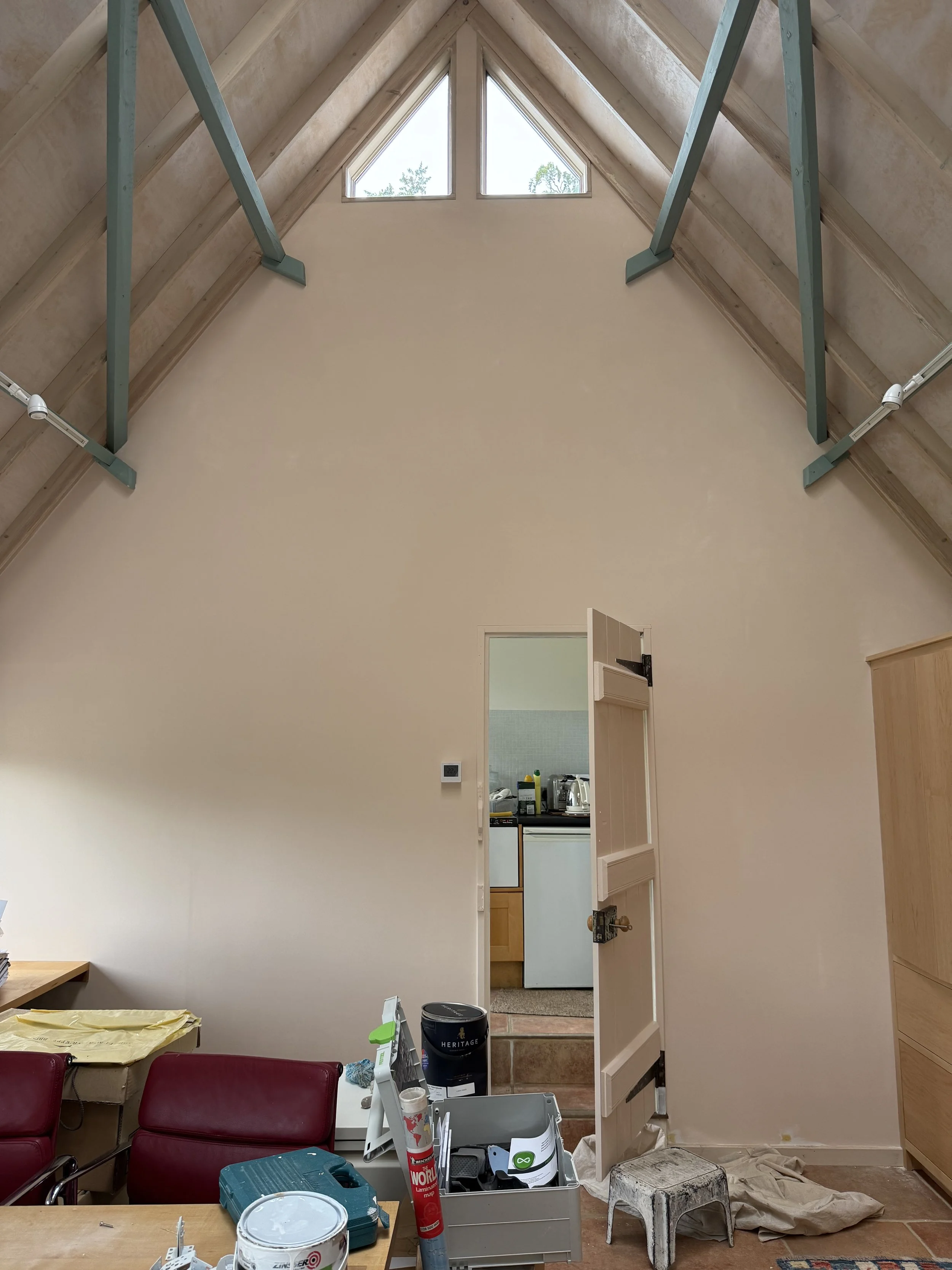 Interior view of a room with a high sloped ceiling, visible beams, and a triangular window at the peak. The door to another room is open, revealing a small kitchen. There are various household items, tools, and furniture, indicating ongoing home reno