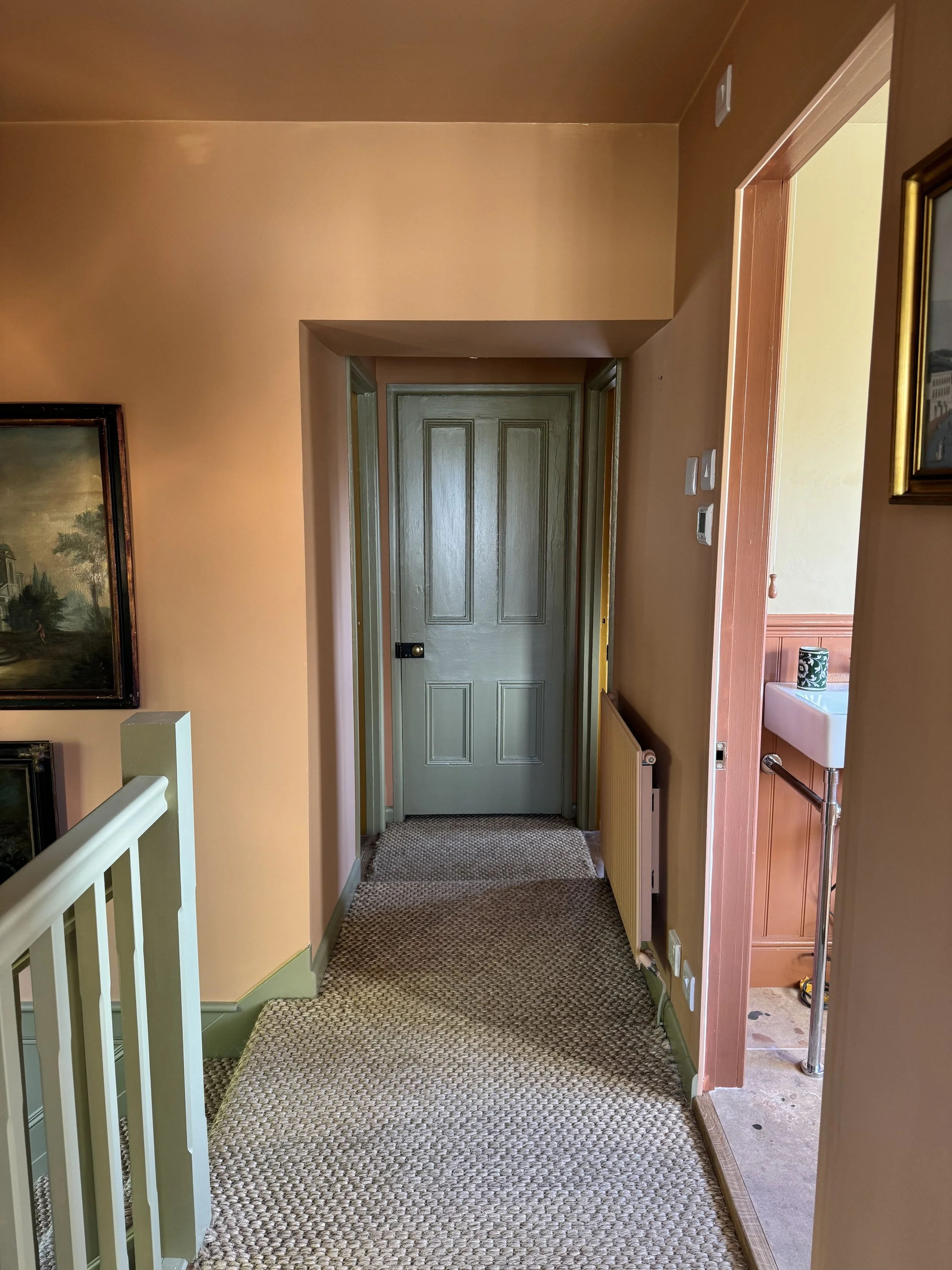 Hallway with beige painted walls, a closed gray door at the end, partially visible staircase on the left, a small radiator on the right, and a doorway to a room with a small sink on the right. The floor has a textured beige carpet.