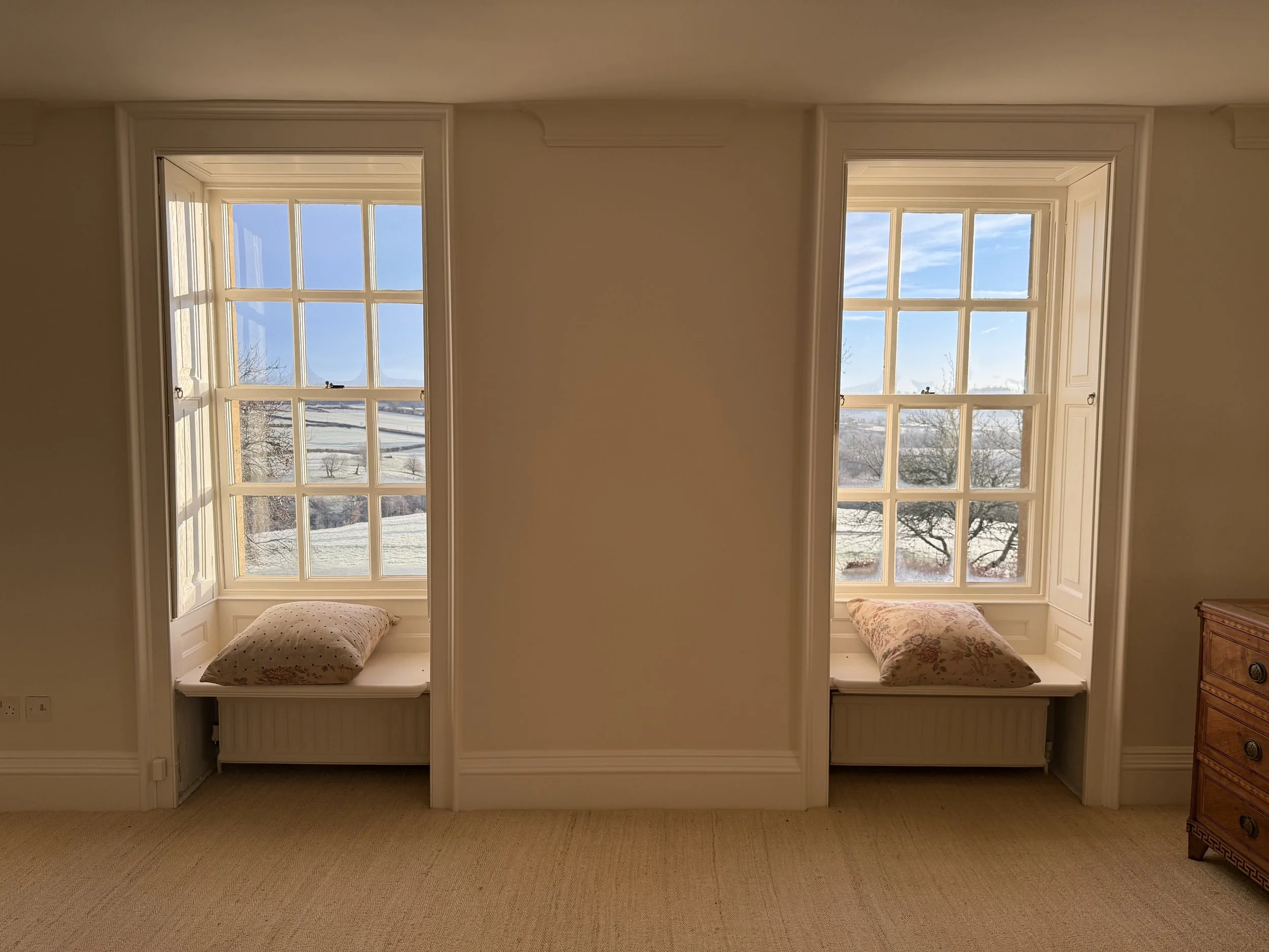 Two large windows with a cozy nook underneath each, featuring pillows and a scenic view of trees and a blue sky outside.