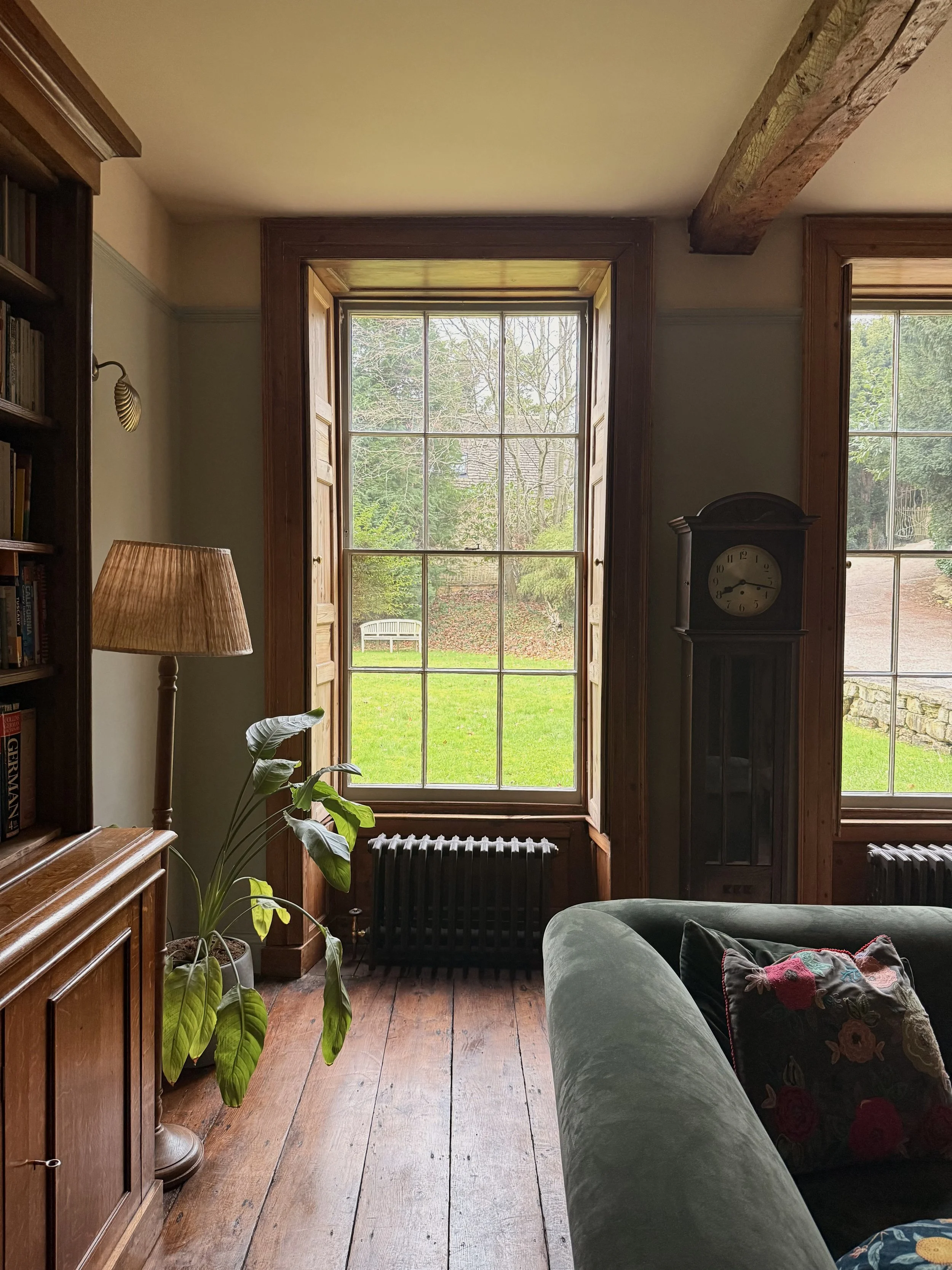 Living room with wooden floors, a large window opening to a green yard, a green velvet sofa with a floral cushion, a vintage grandfather clock, a tall potted plant, a wooden bookcase, and a floor lamp with shades.