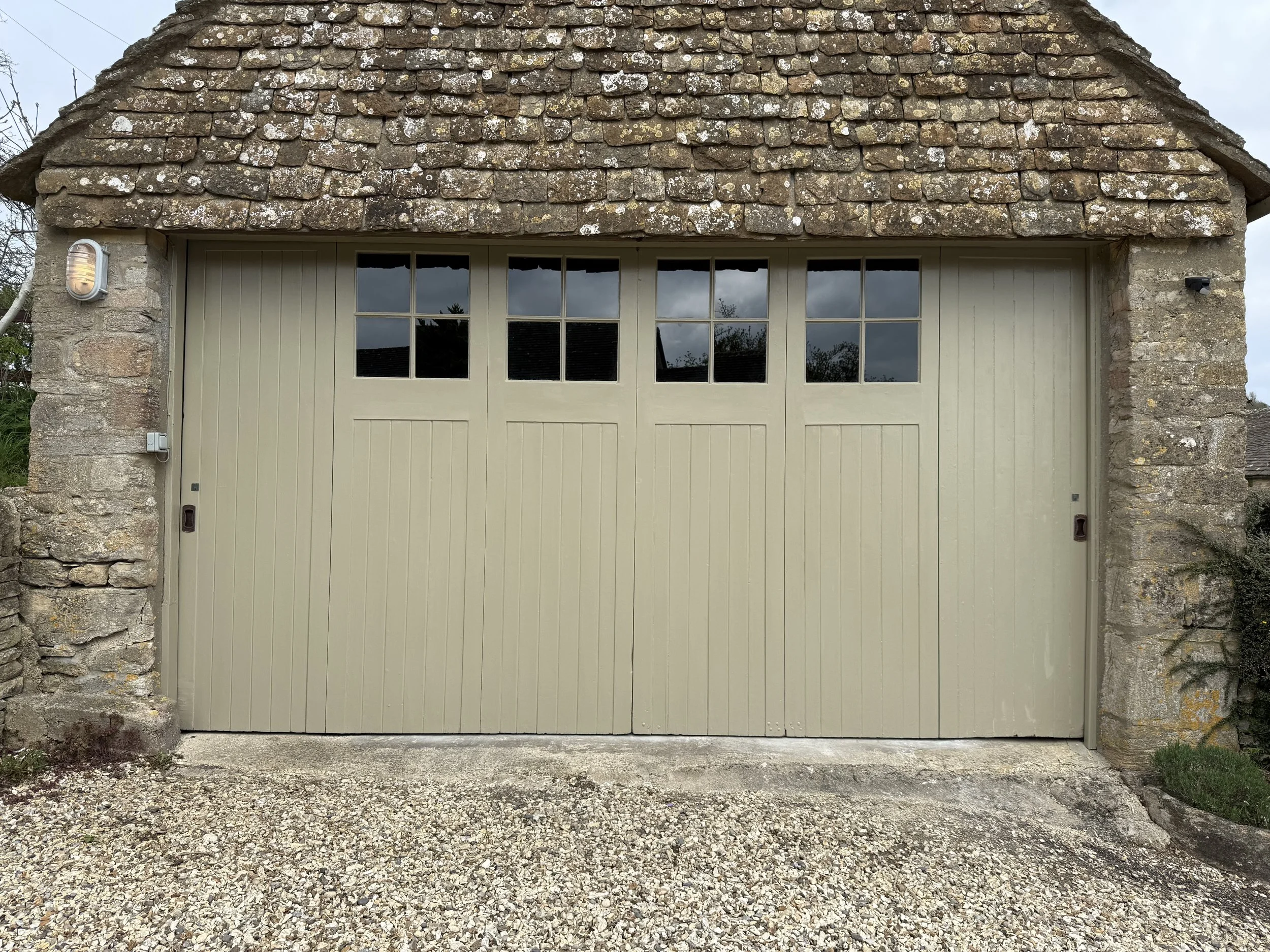 Stone garage with a beige, paneled door and four small window panes at the top, situated under a shingled roof with lichen growth.