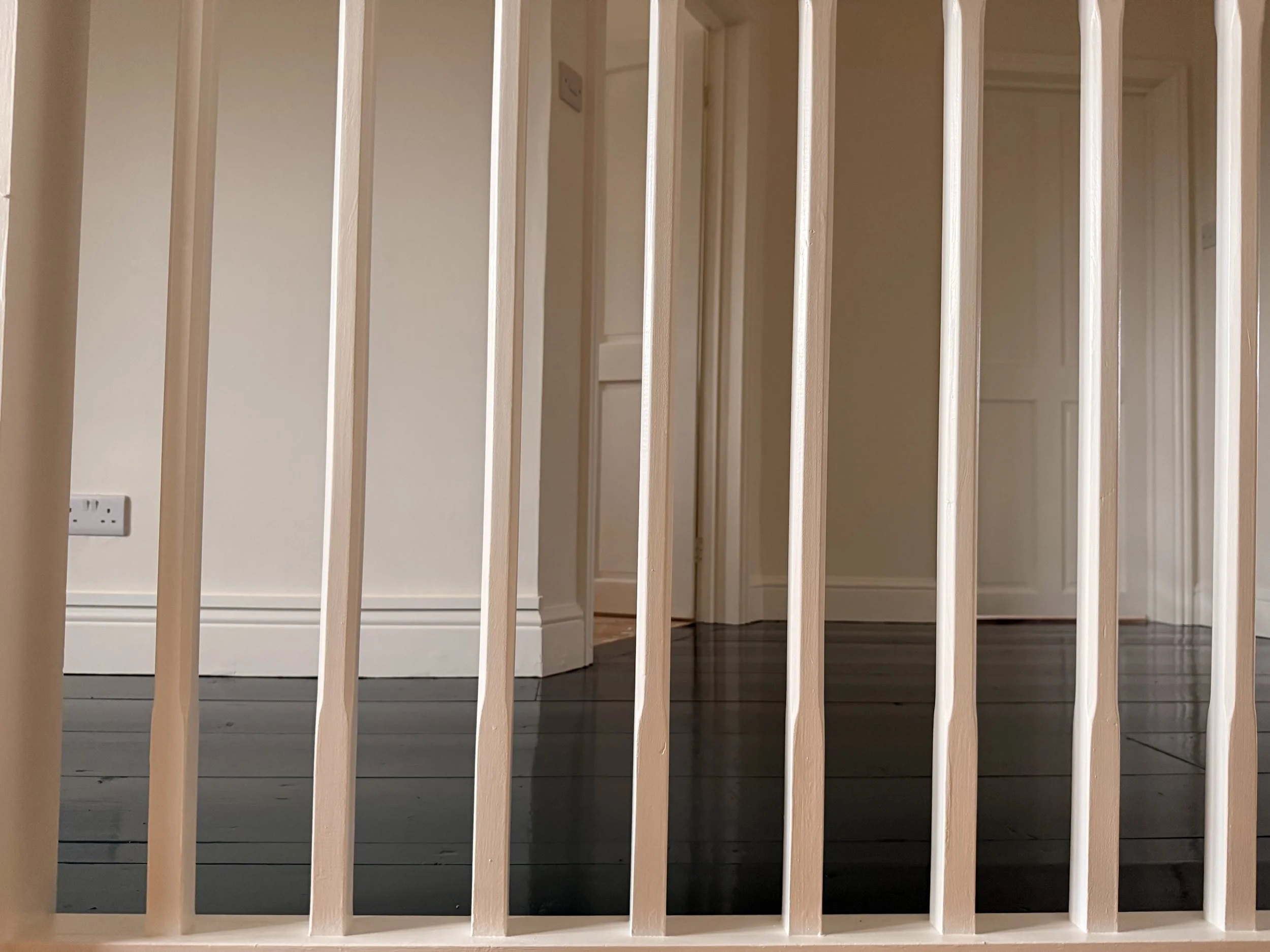Interior view of a room seen through a wooden baby gate with vertical bars. The room has beige walls, white baseboards, and dark-colored wooden flooring. There is a visible electrical outlet on the wall to the left.