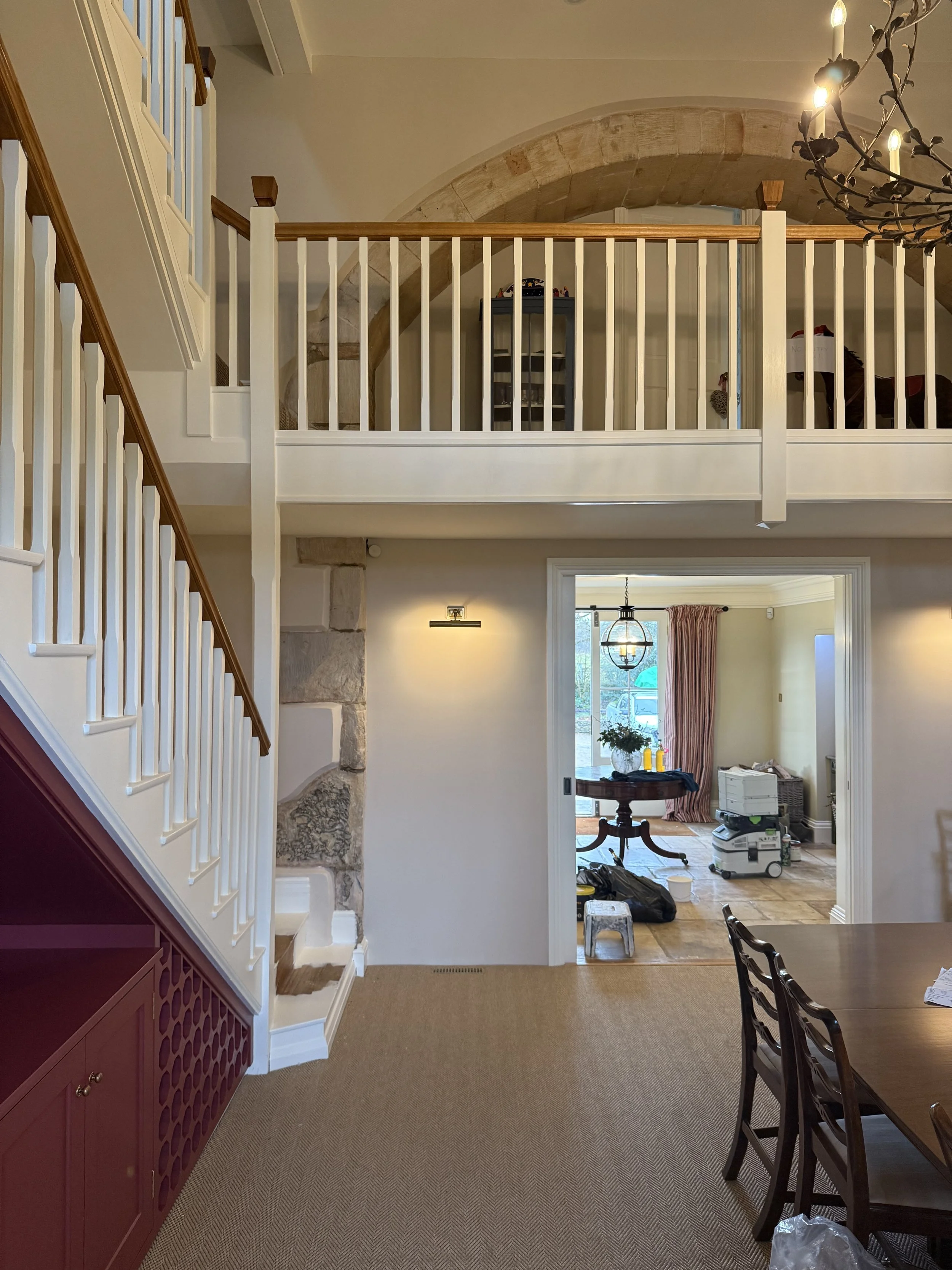 A view of an interior room showing a staircase, a doorway, and a dining table with chairs. The staircase has white railings and wooden handrails, and the room beyond the doorway contains cleaning supplies and a window with curtains.