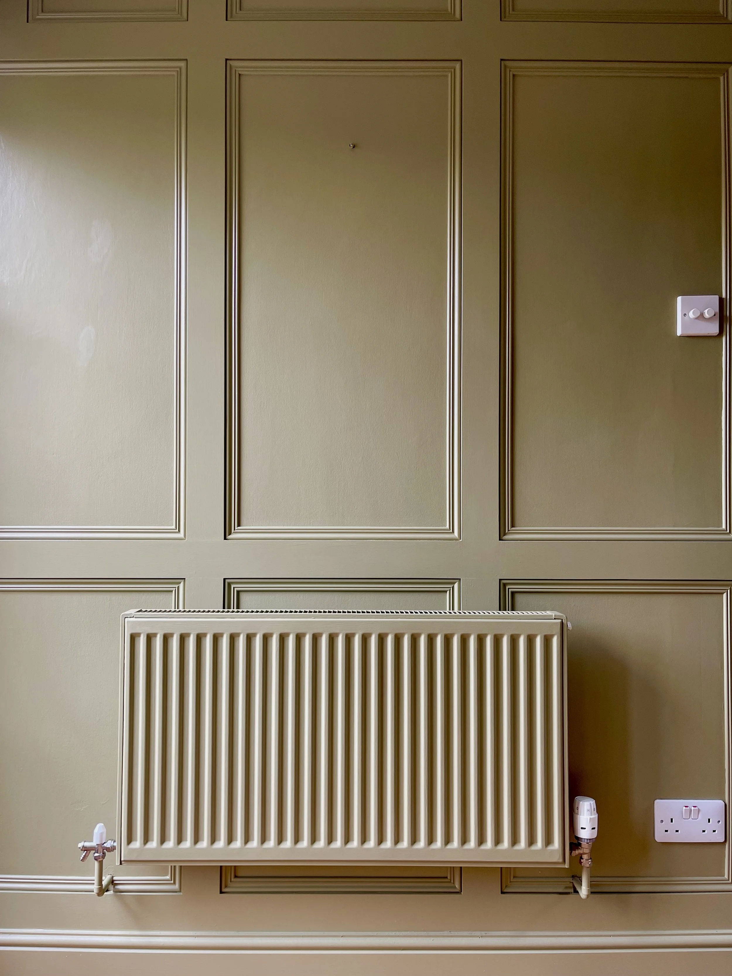 Cream-colored wall with decorative paneling, radiator, electrical outlet, and thermostat.
