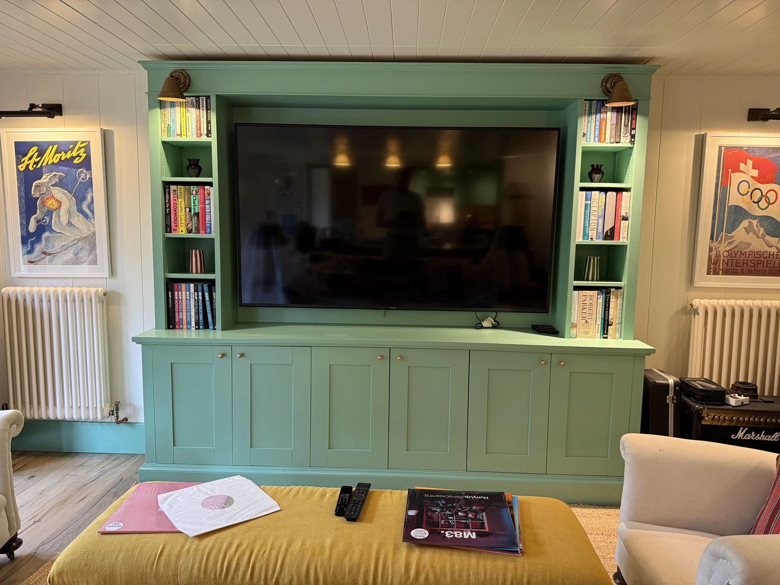 Living room with a large black flat-screen TV on a green entertainment unit with bookshelves on either side, surrounded by framed posters and a radiator, with a yellow ottoman with magazines and remote controls in front.
