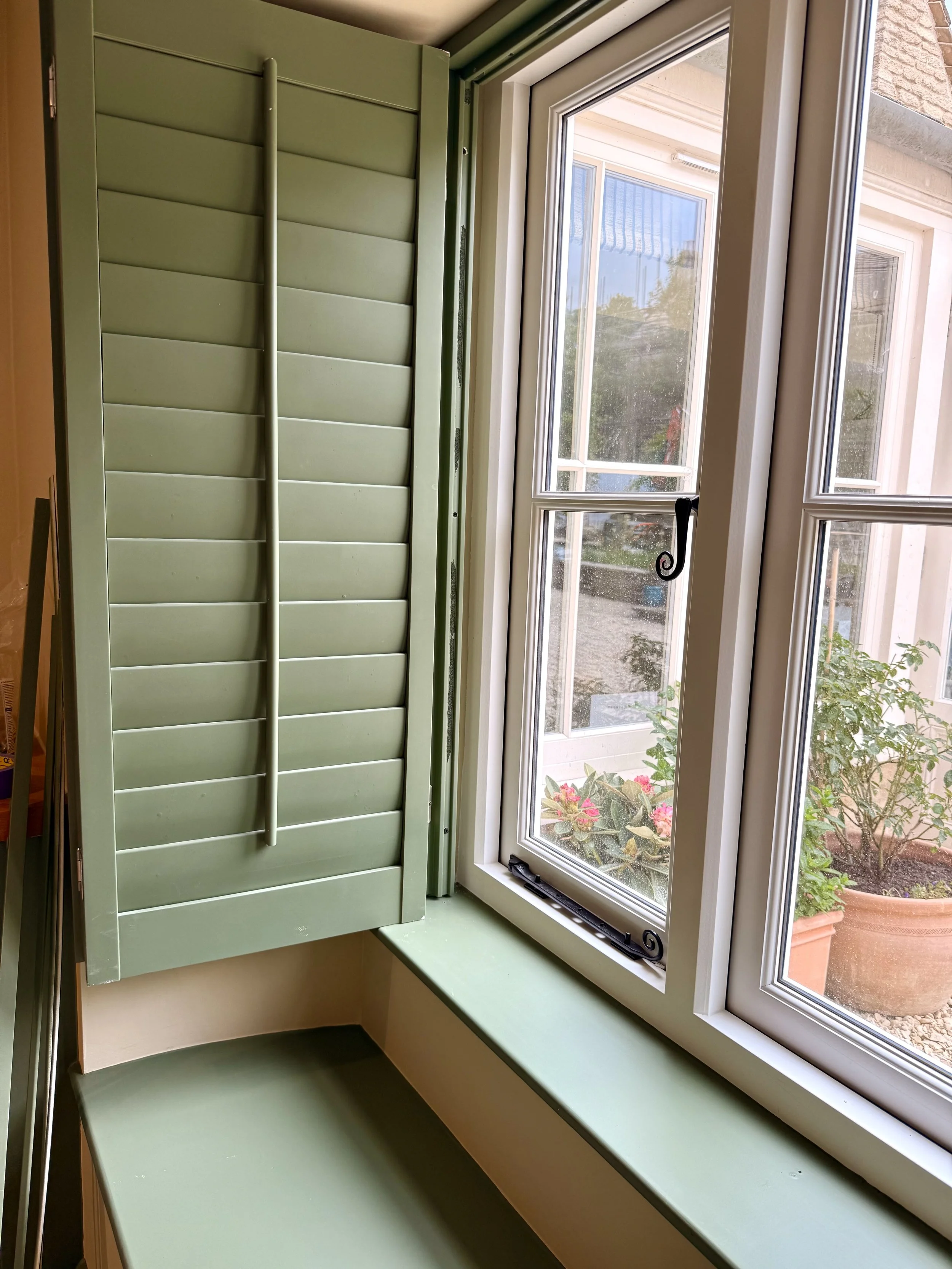 Green window shutter partially open next to a white-framed window with a view of plants outside.