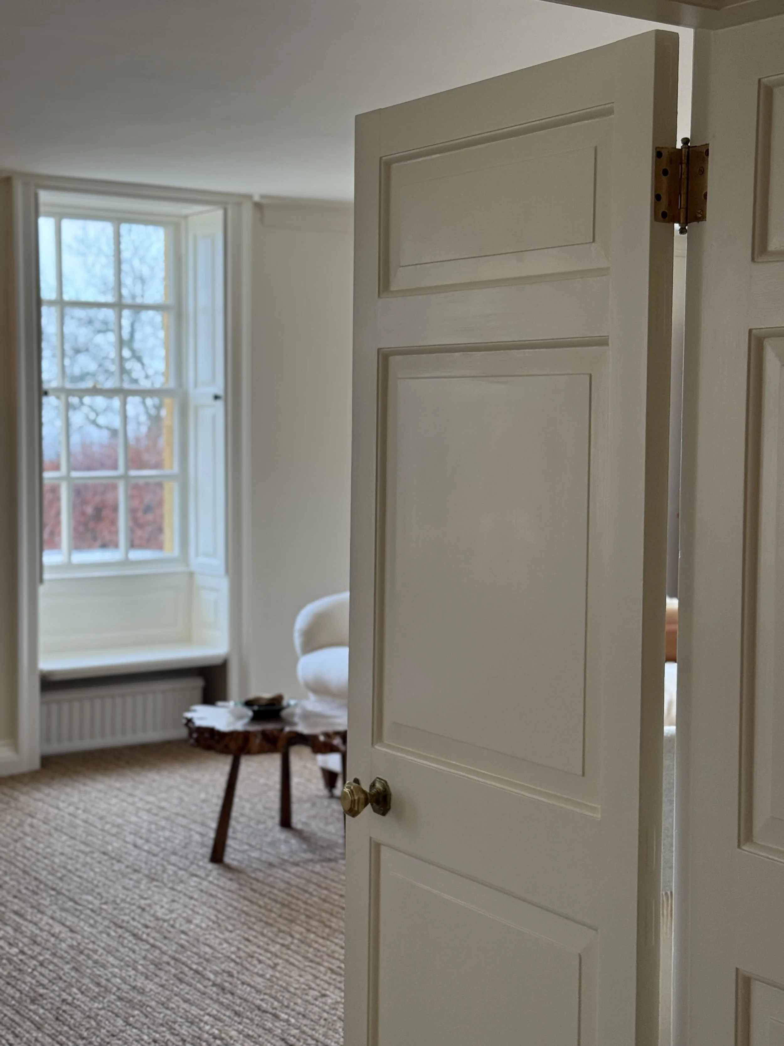 Interior view of a room with a partially opened white door, a window with multiple panes, and a table with clutter, in a cozy setting.