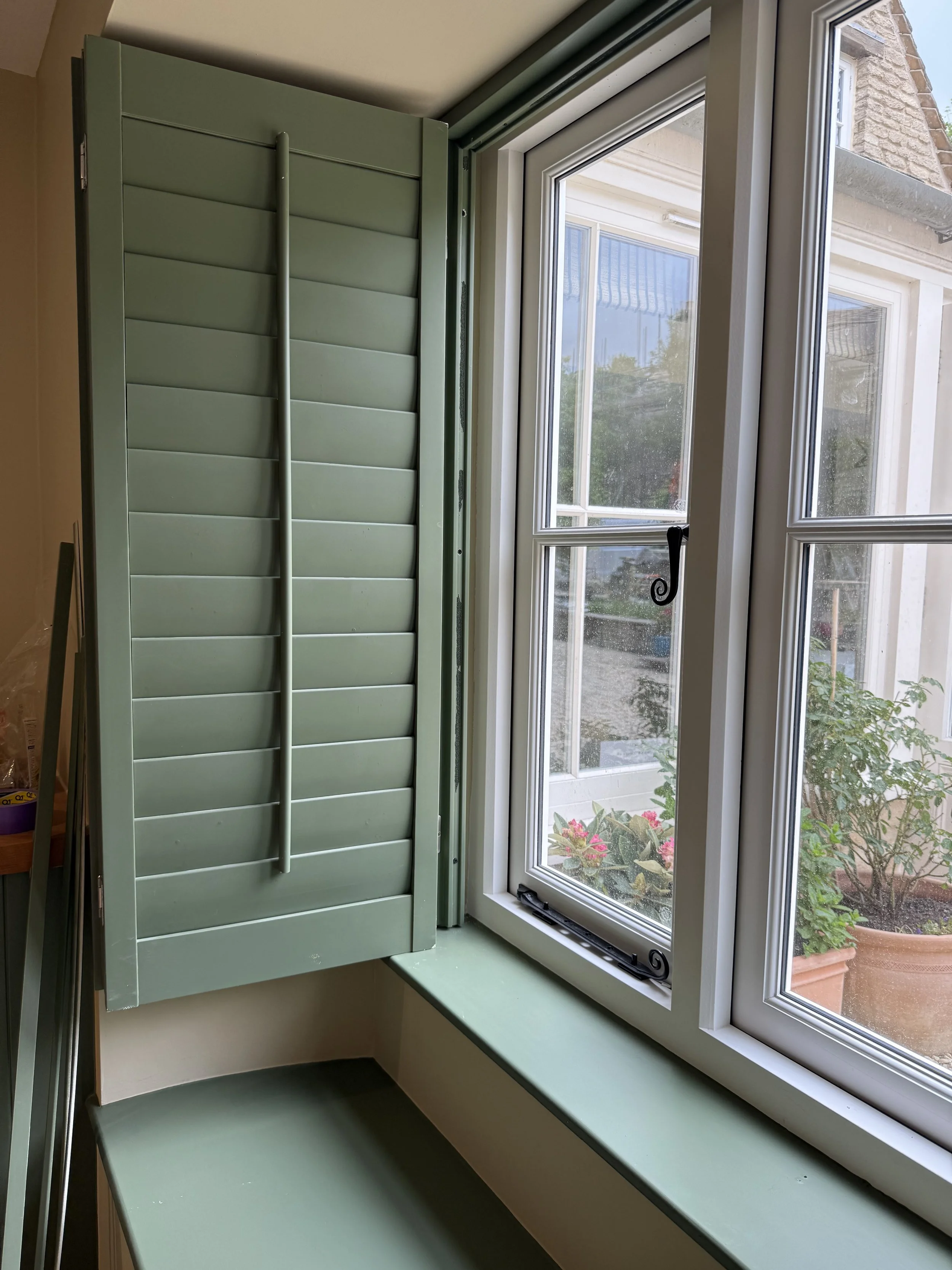 View from inside a house showing a window with green shutters partially open, revealing an outdoor garden with potted plants.