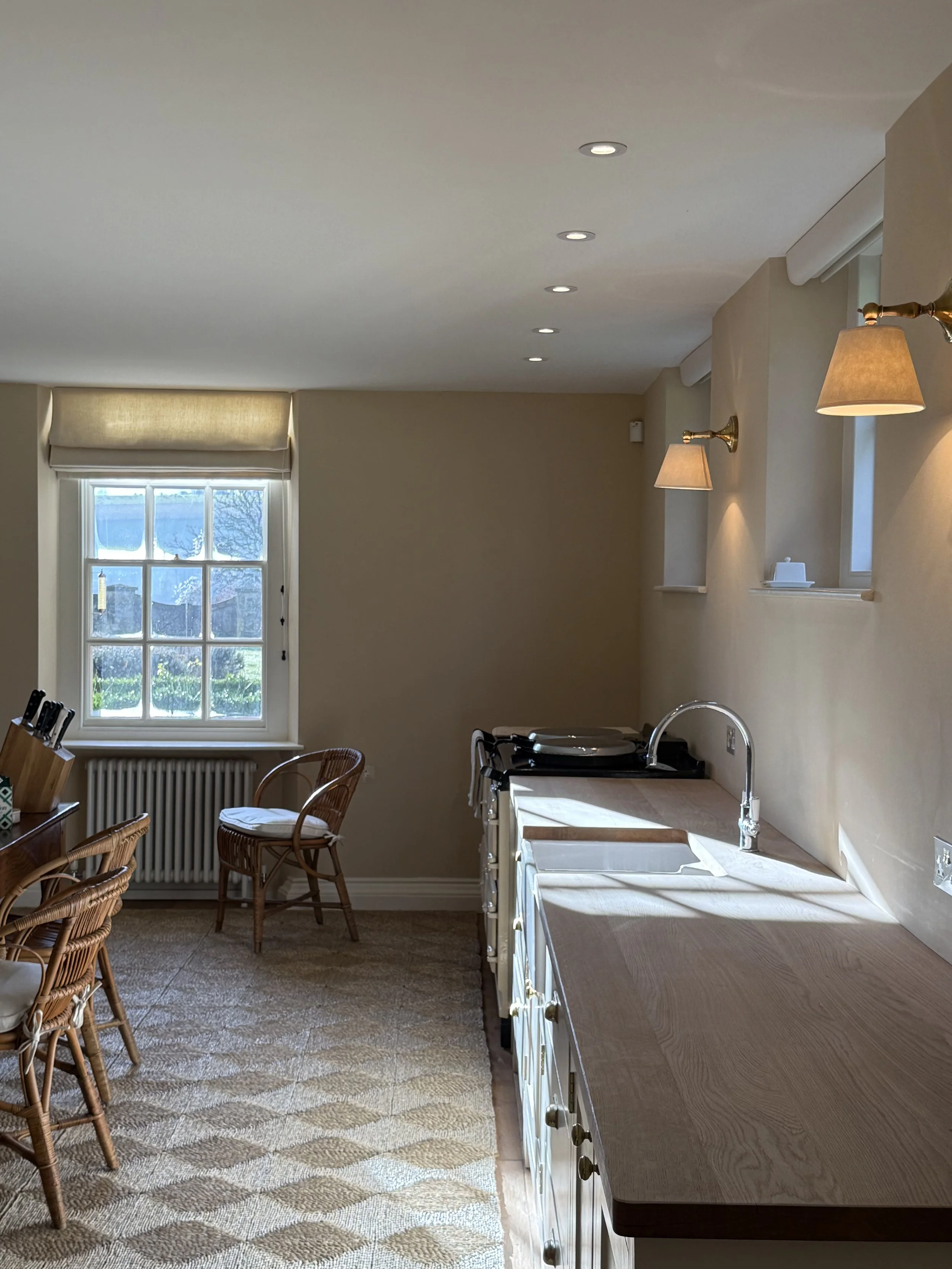 Sunlit kitchen with wooden countertops, white cabinets, a stainless steel sink, and wall-mounted lamps, with a window showing greenery outside.