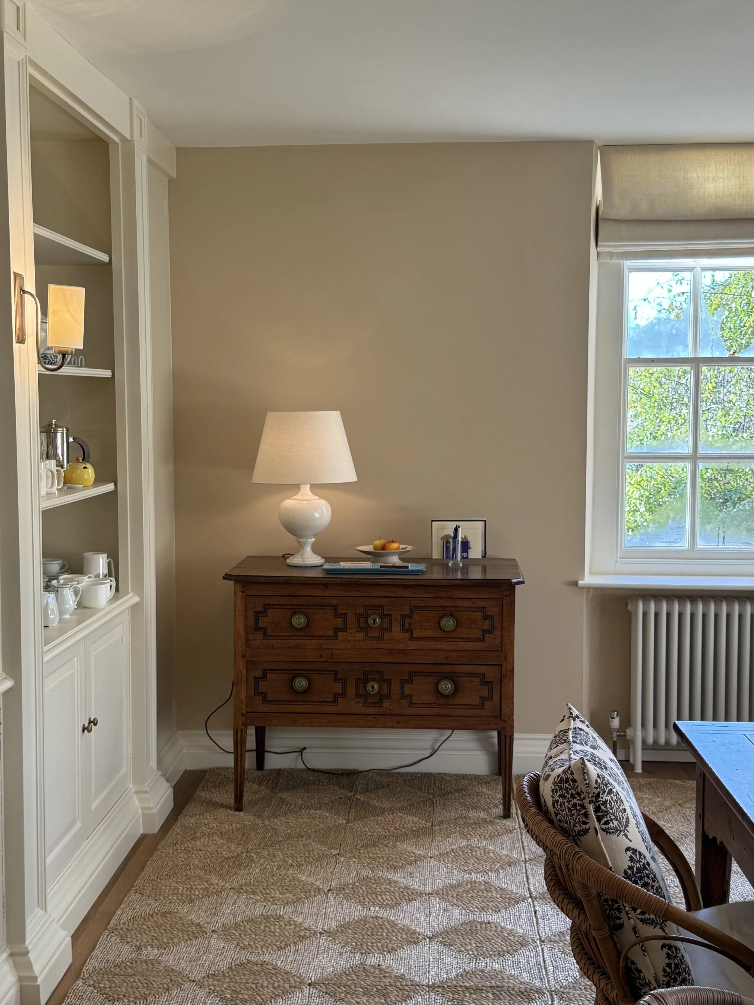 Interior of a well-lit room with a wooden dresser, a white table lamp, a bowl of fruit, a framed photo, a large window with a view of trees, a patterned rug, a woven chair with black and white cushions, and a built-in white shelving unit with dishes.