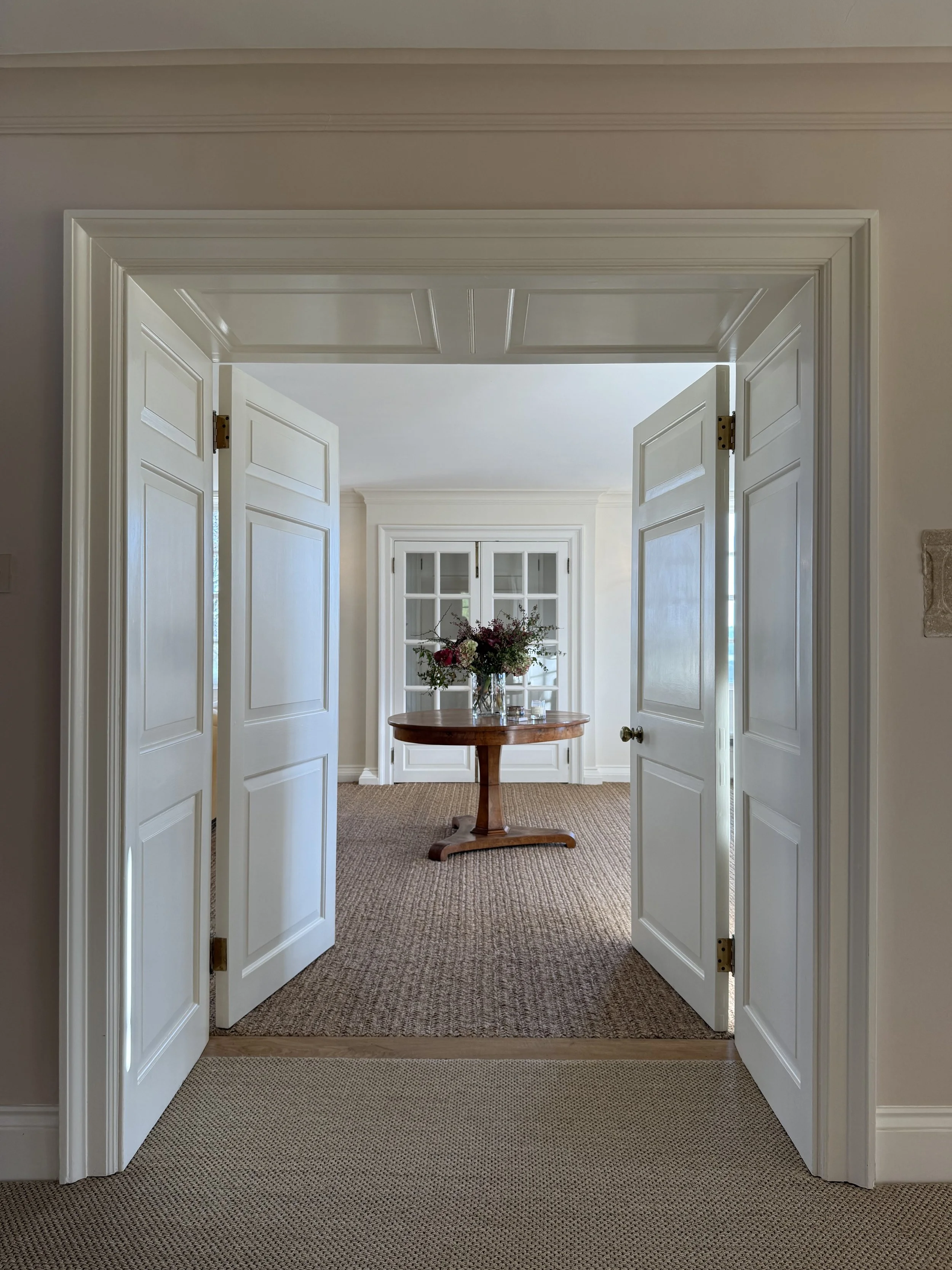 Open double white interior doors leading to a room with a round wooden table and a vase of flowers, white window cabinets, and beige carpet.