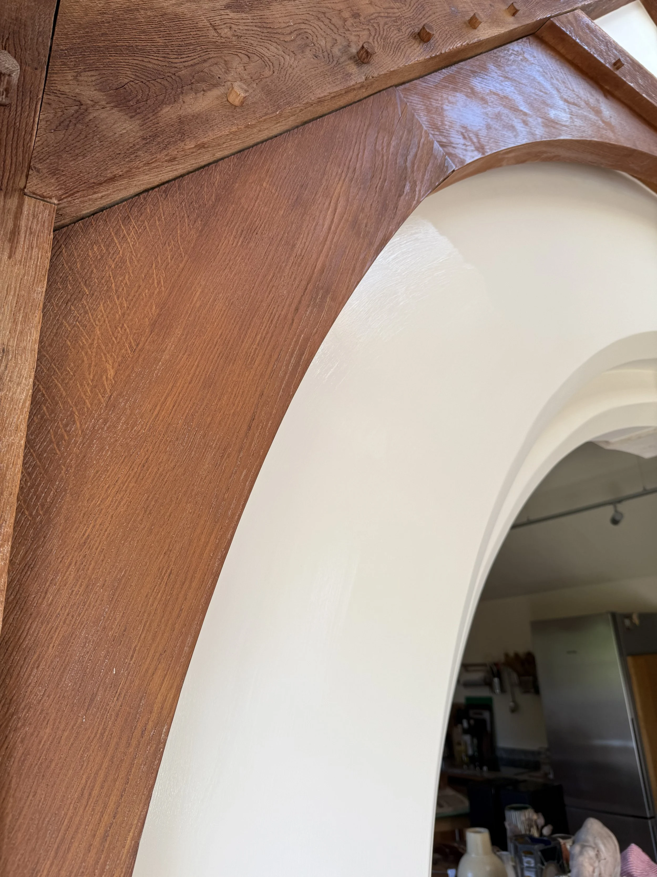 Close-up of a wooden archway with visible wood grain, adjacent to a white wall and photo of a kitchen in the background.