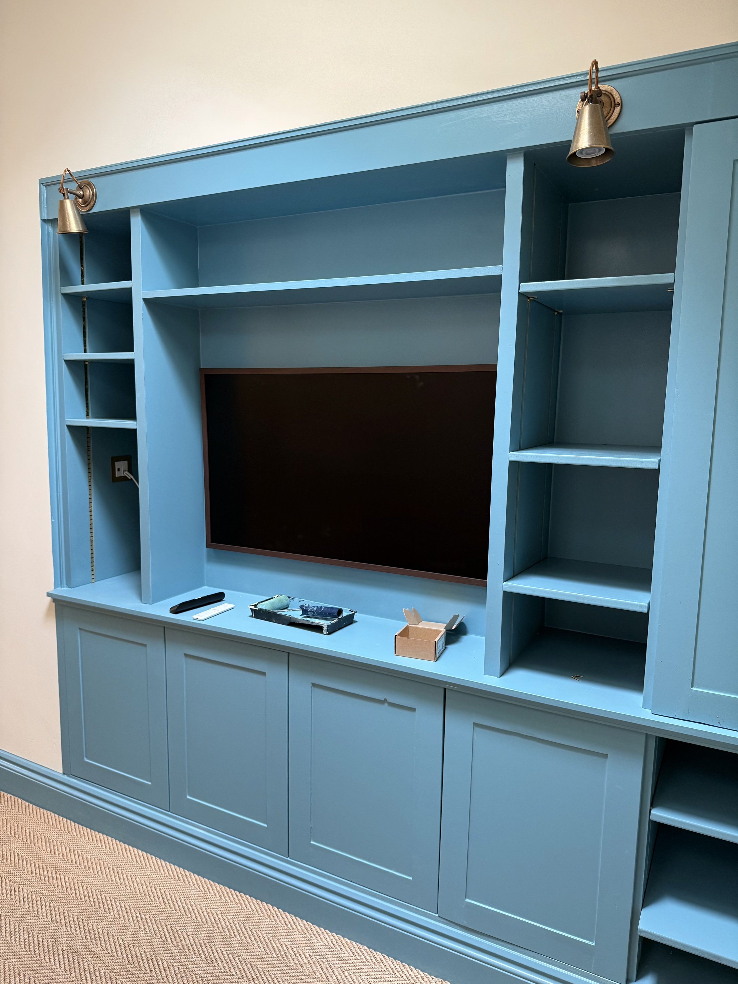 Blue built-in entertainment center with a large blank TV space, shelves, and adjustable lights, on a beige carpeted floor.