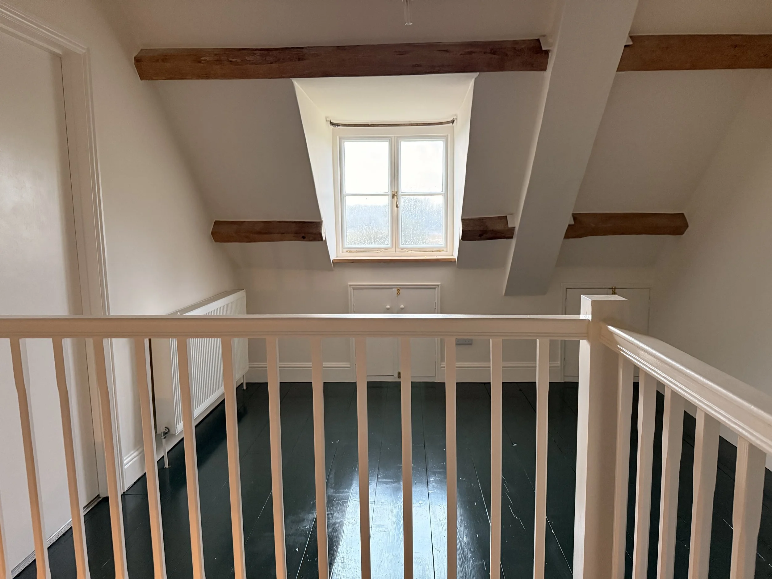 View of an attic room with a window, exposed wooden beams, a radiator, and white walls, visible from behind a white railing.