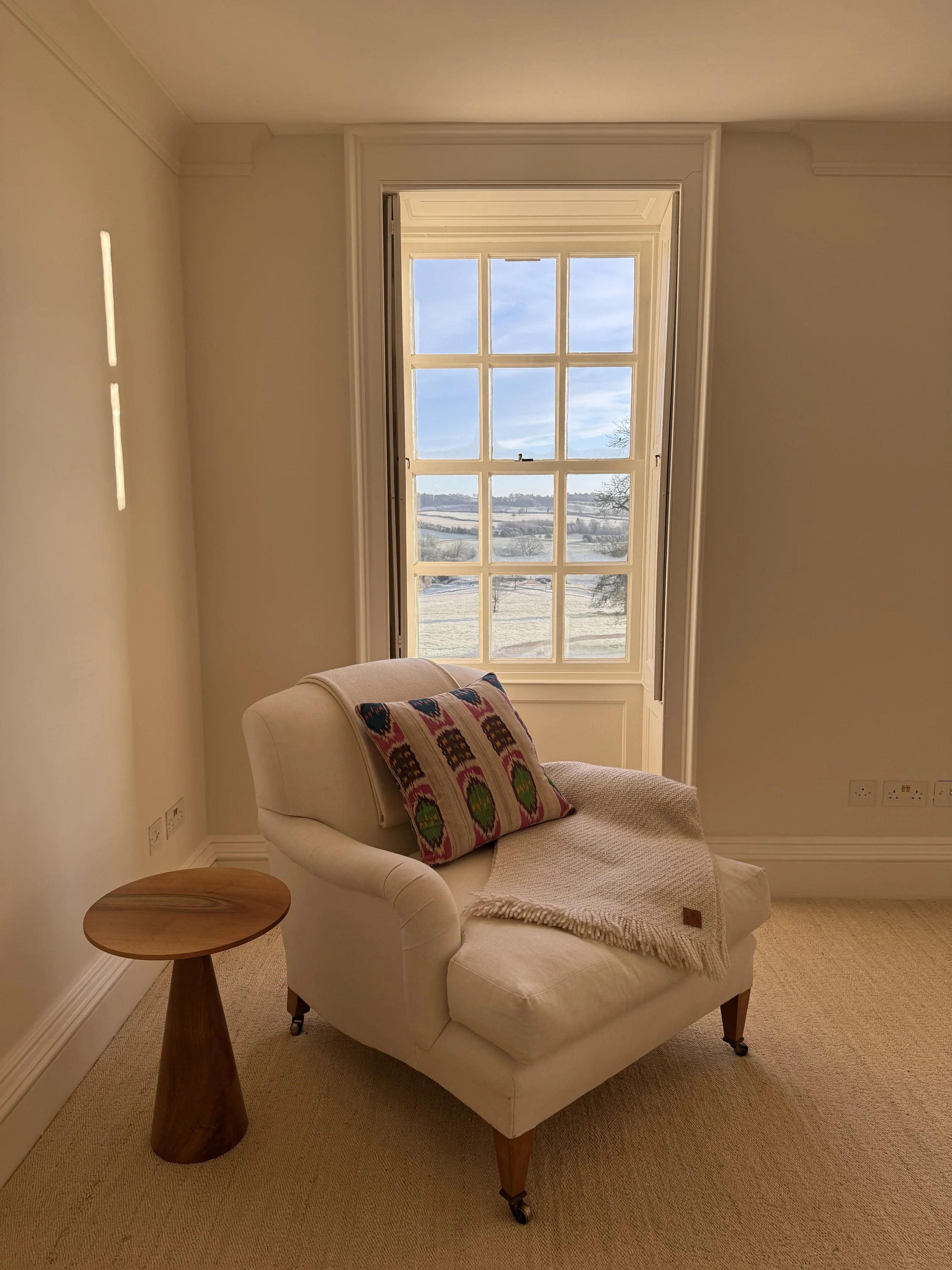 A cozy corner in a room with a beige armchair, a wooden side table, a colorful embroidered pillow, a cream throw blanket, and a large window showing a snowy landscape with blue sky and scattered clouds.