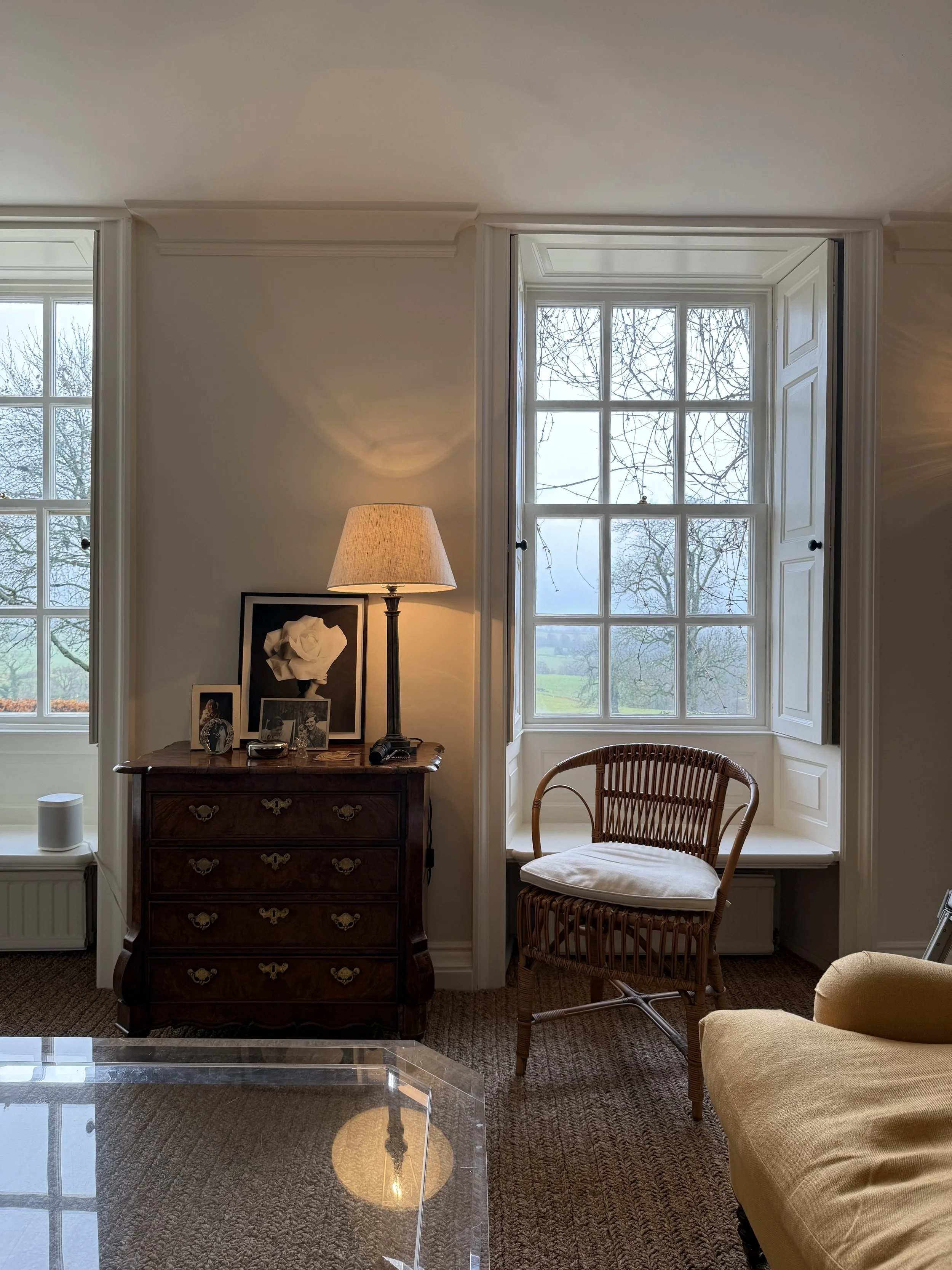 A cozy living room corner with a wooden dresser topped with framed photos and a lamp. There are large windows with open shutters, revealing a view of leafless trees and a cloudy sky outside. A woven chair with a cushion is situated next to the dresse