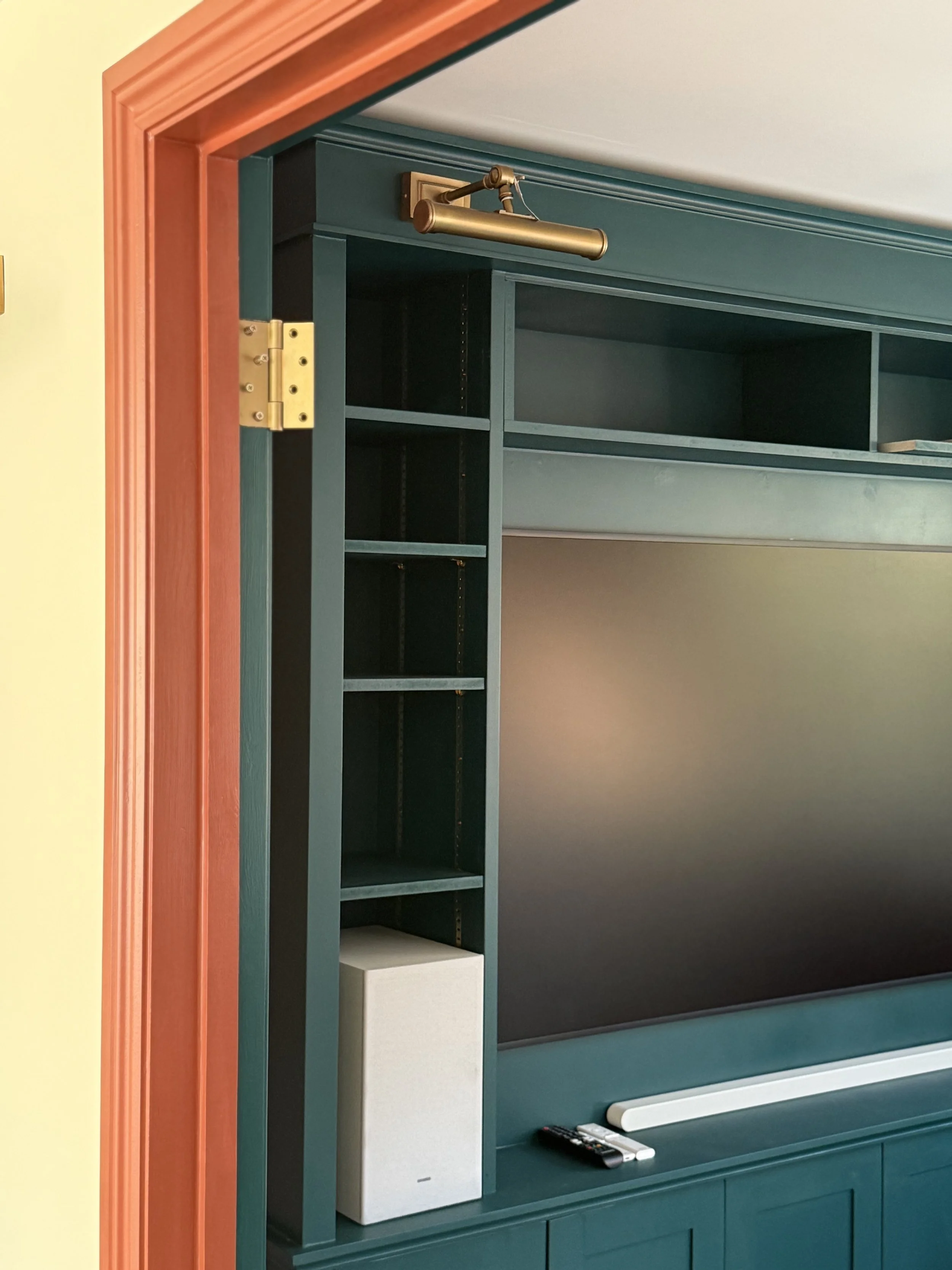 Close-up of a teal built-in bookshelf with an open section and a white speaker, mounted on a teal wall with a dark-colored TV or monitor below, and a remote control and white electronic device in front.