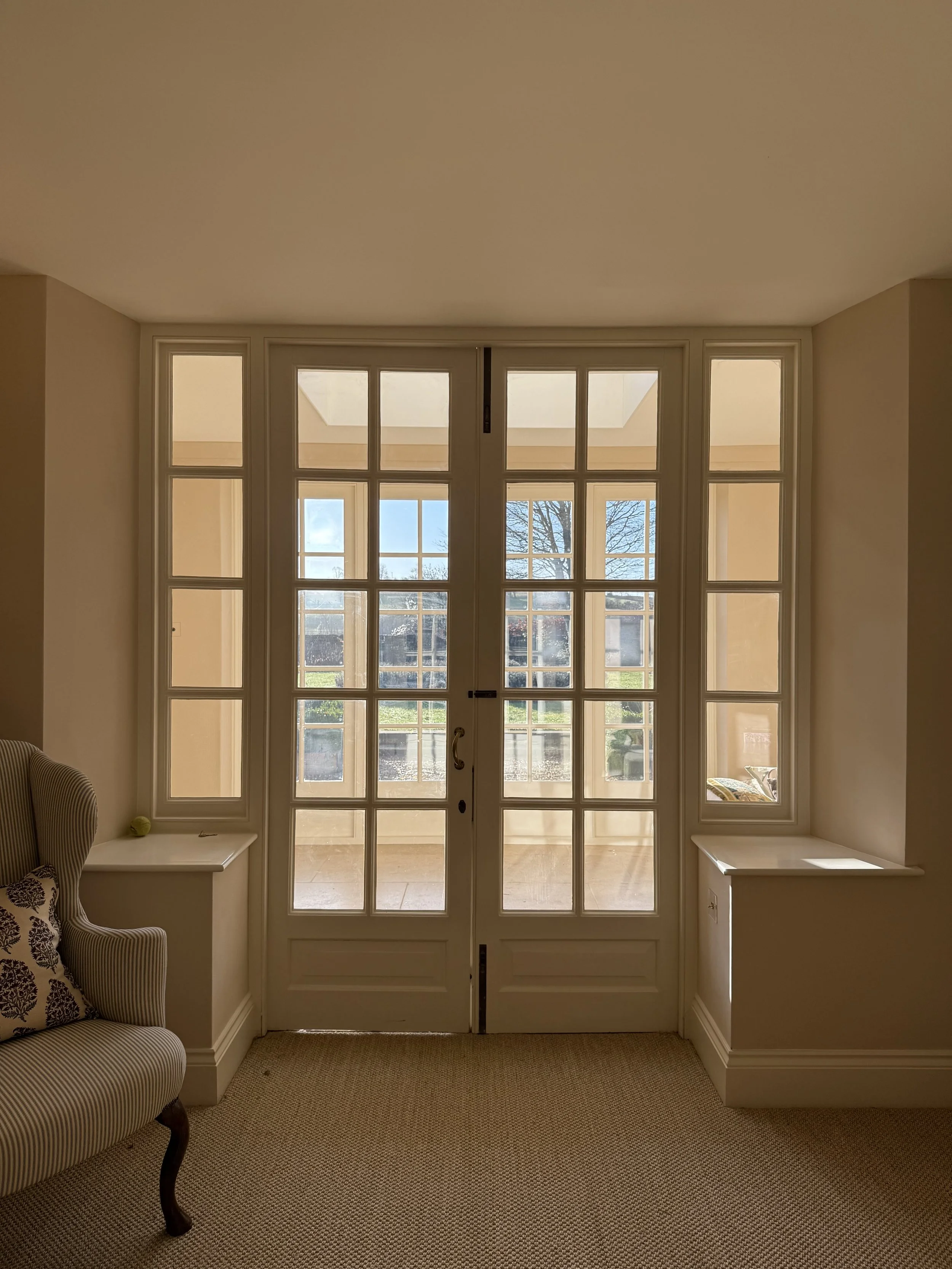 Interior view of a room with large glass double doors leading to a patio, with windows on either side showing an outdoor scene with trees and a partly cloudy sky.