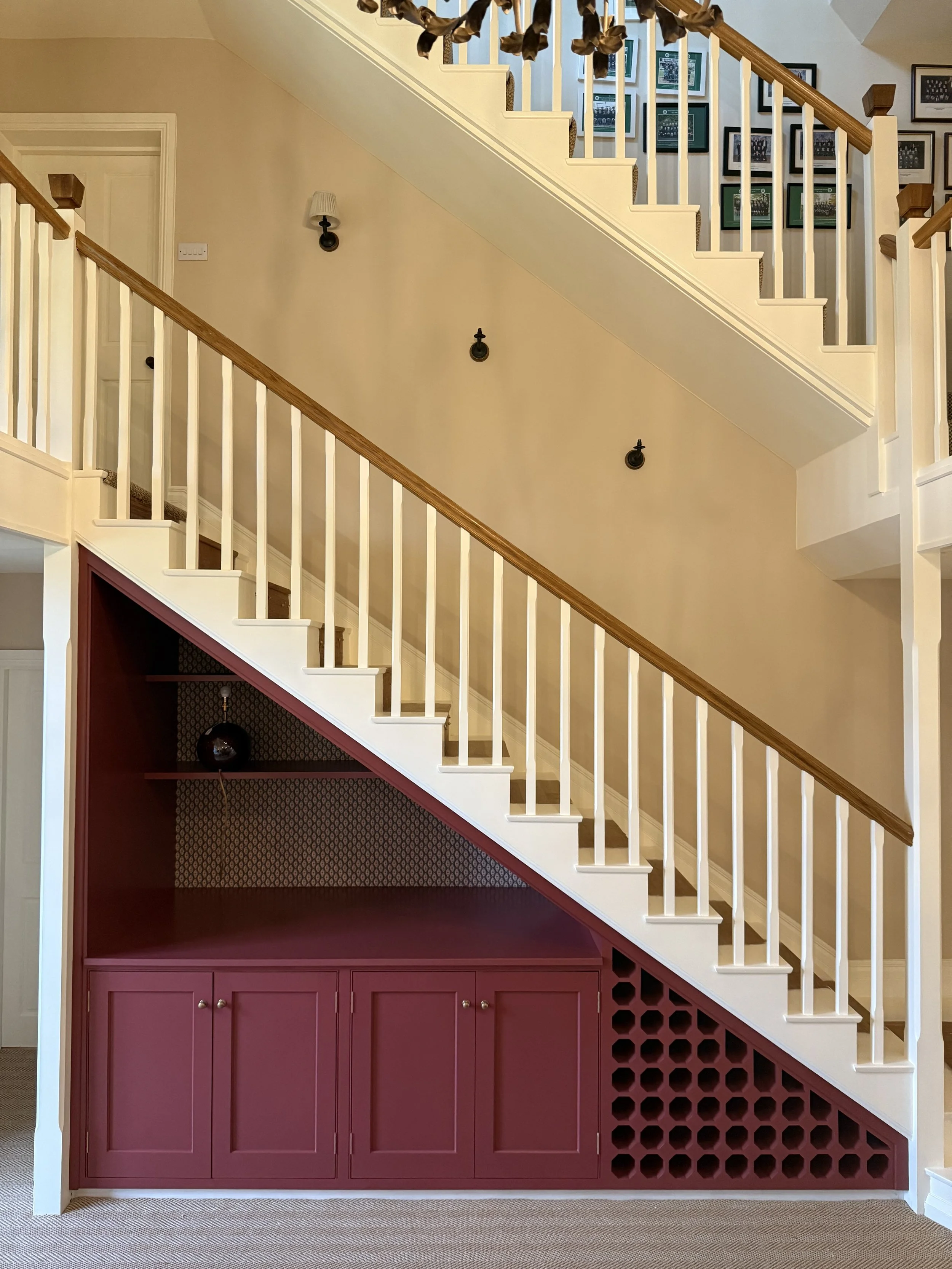 Interior view of a staircase with a built-in burgundy-colored storage cabinet and circular holes, white railings, framed photos on the wall, and a ceiling light fixture.