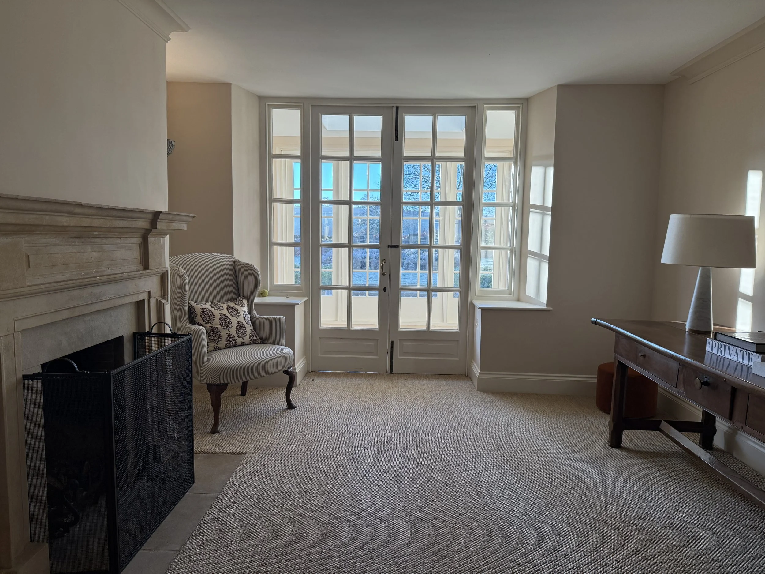 Living room with beige carpet, a fireplace on the left, a striped armchair with a patterned pillow near the window, a side table, and a wooden console table with a white lamp on the right. Large windows and glass doors with white frames let in natura