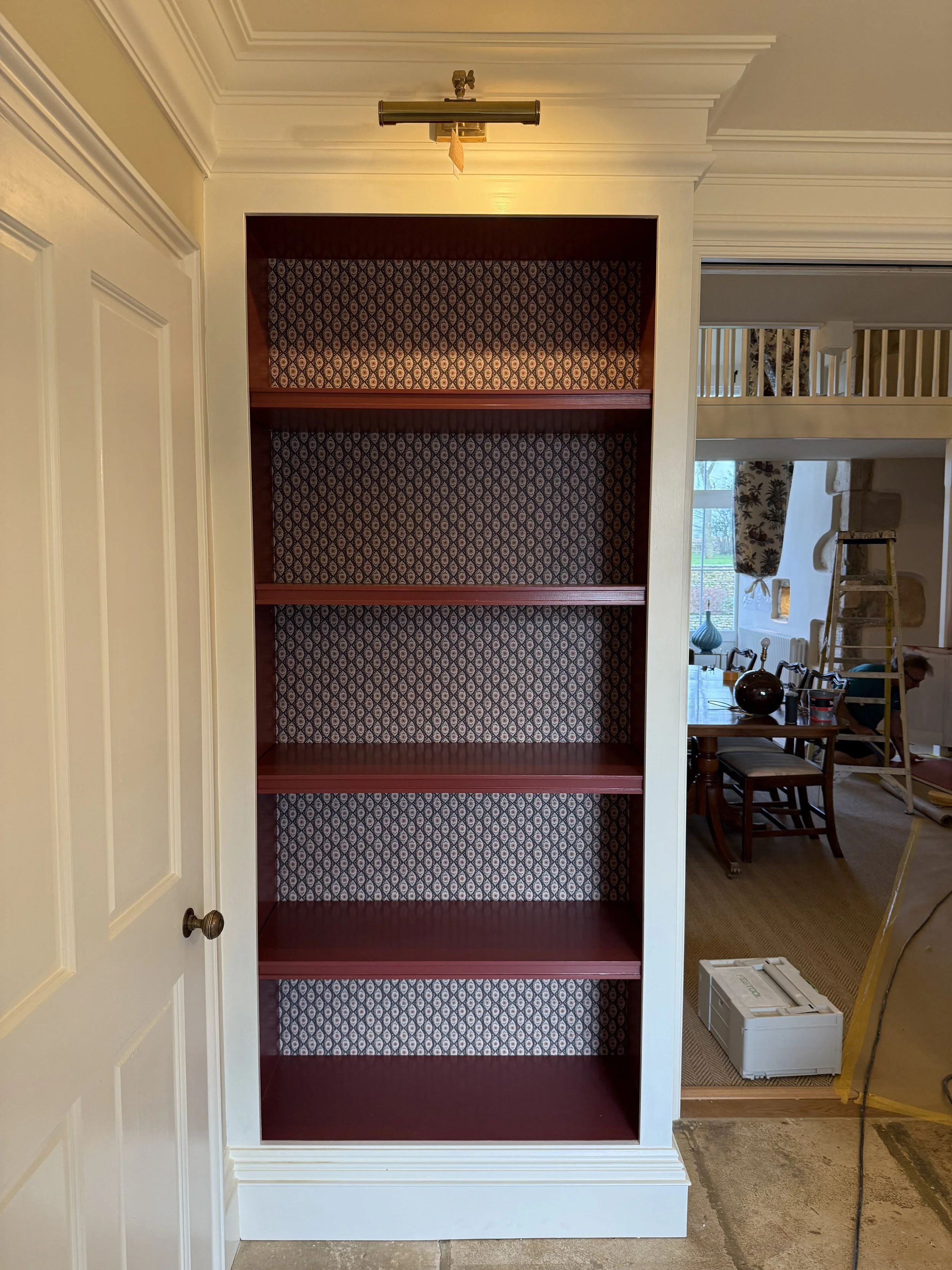 Empty wooden bookshelf with patterned wallpaper backing in a room under renovation.