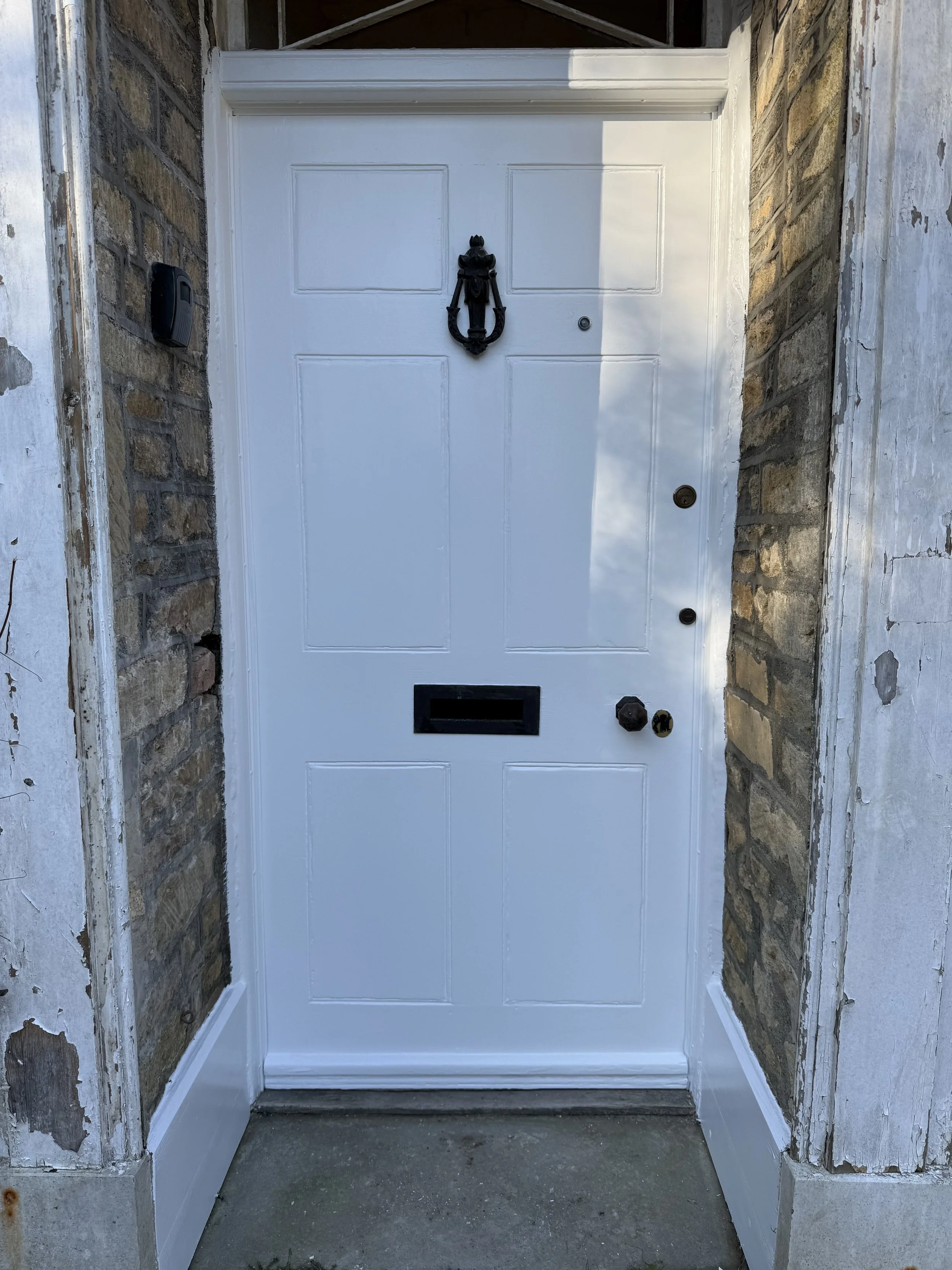 White front door with a black decorative door knocker, a black mail slot, and multiple locks. The door is framed by brick and weathered white siding.