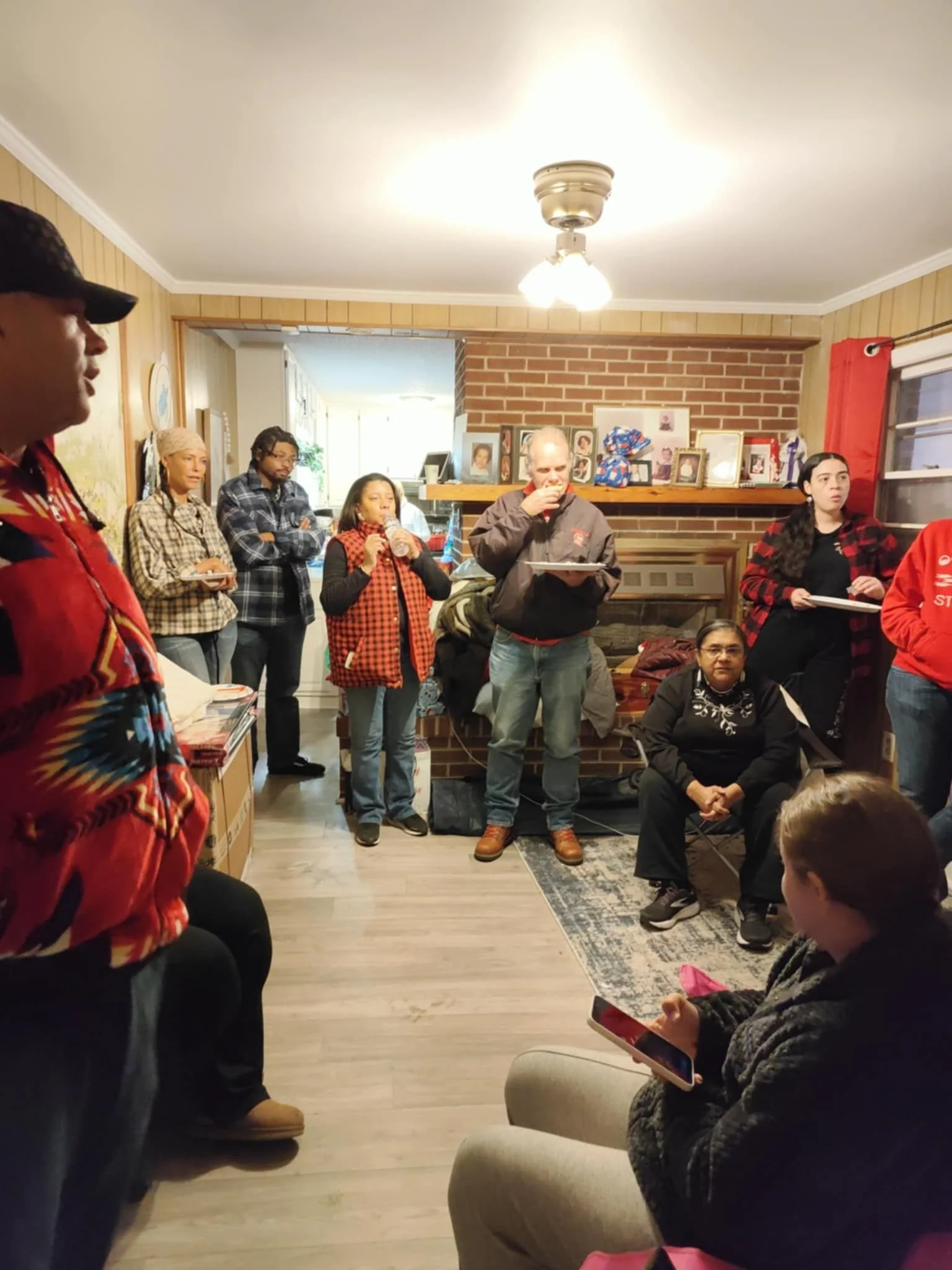 A group of people gathered in a living room, with some standing and some sitting, engaging in a social event. The room has a brick fireplace with family photos on the mantel, a ceiling light, and a window with red curtains.