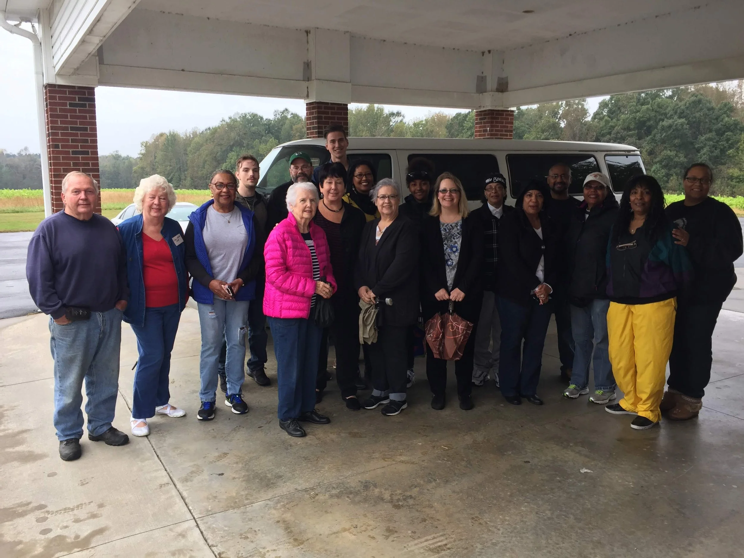 A group of diverse people standing together under a porch with a scenic rural background, smiling for the photo.
