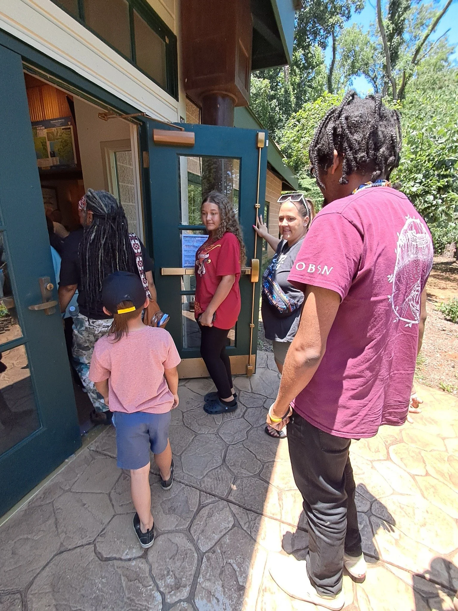 A group of people standing in line at a door to enter a building on a sunny day, with trees and a blue sky in the background.