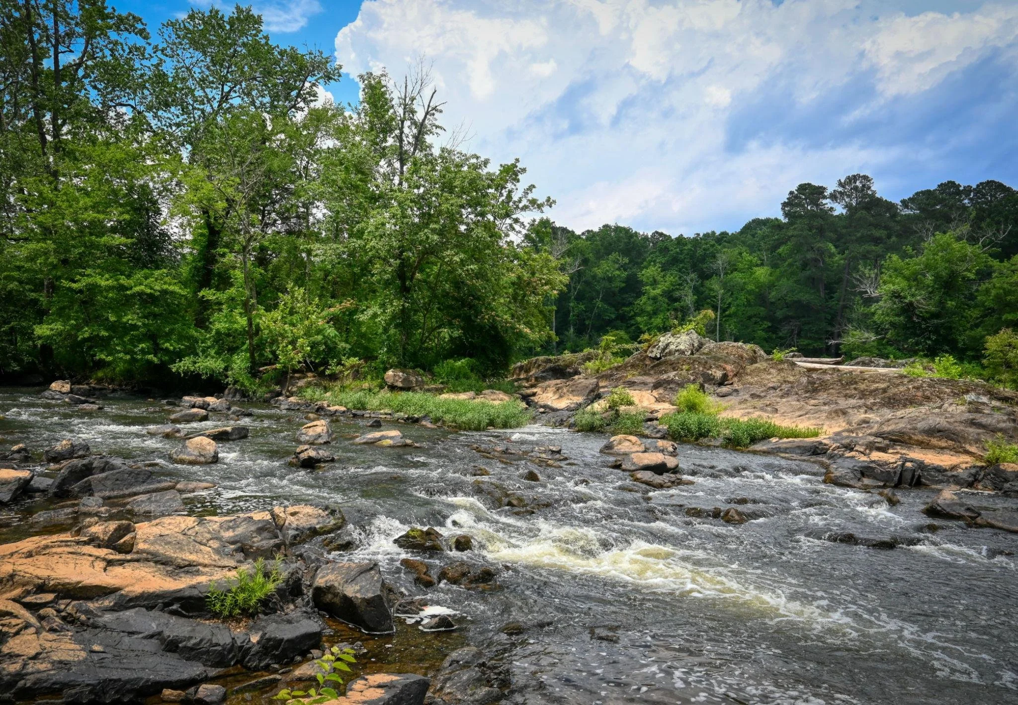 A scenic view of a flowing river with rocks and surrounding green trees under a partly cloudy sky.