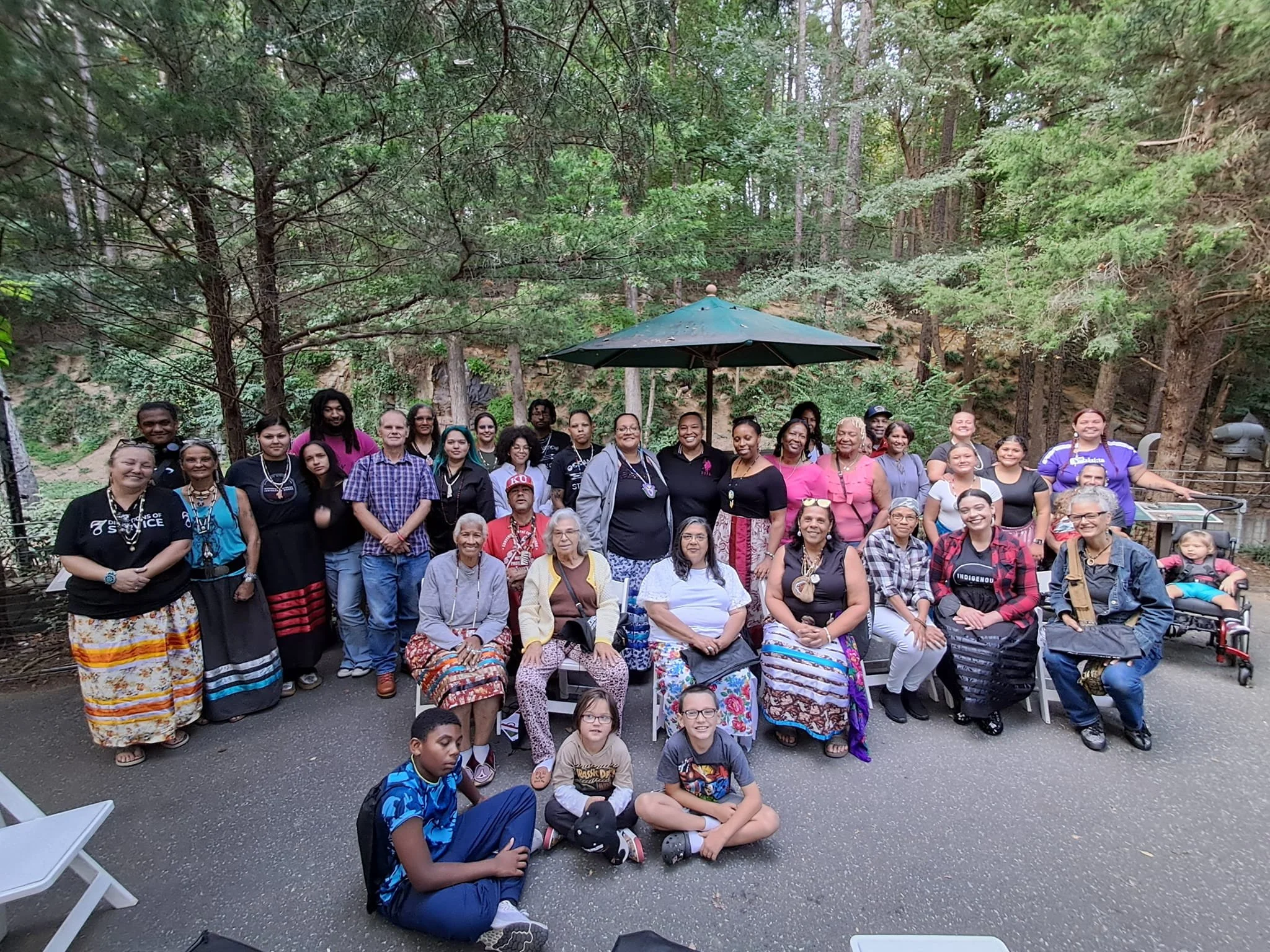 A large group of people, diverse in age and ethnicity, gathered outdoors in a wooded area for a group photo, with some sitting and others standing in front of dense greenery and tall trees.