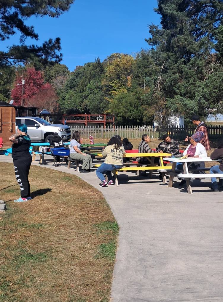Group of people sitting at picnic tables outdoors on a sunny day with trees and a blue sky in the background.