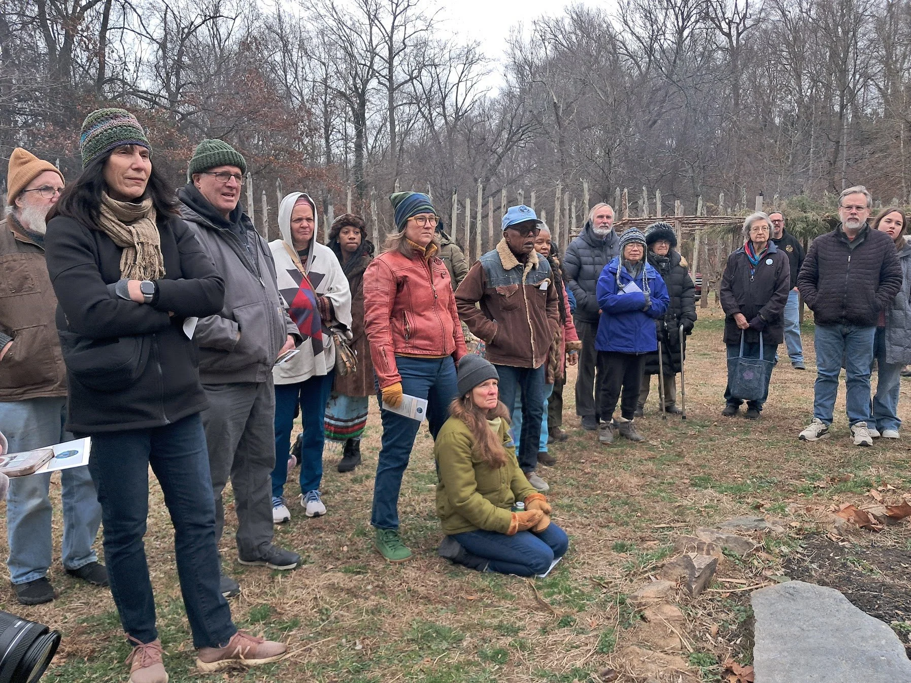Group of people gathered outdoors in a wooded area on a cloudy day, some standing and one kneeling in the foreground, looking attentive.