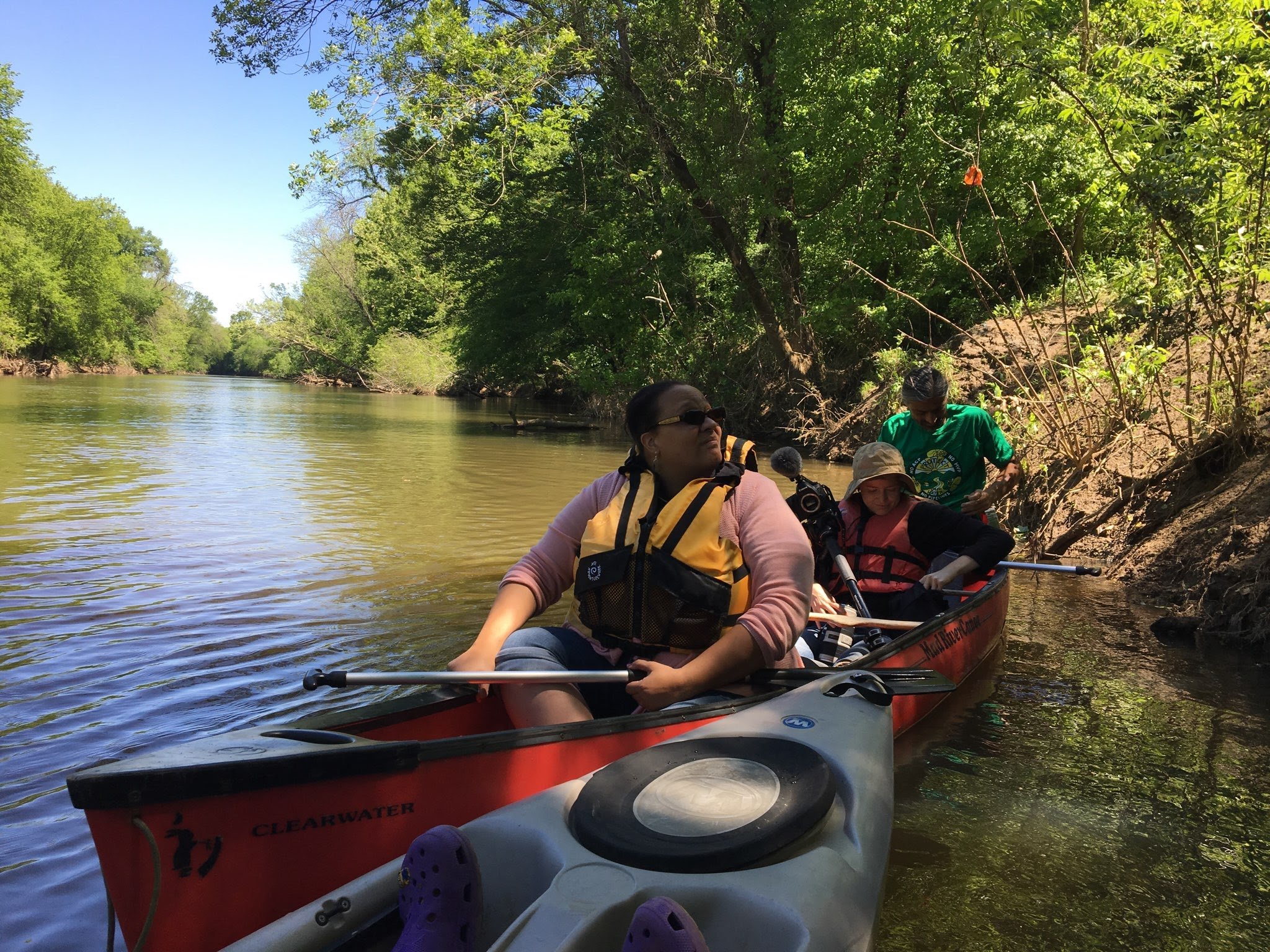 People kayaking on a wide, calm river surrounded by dense green trees under a clear blue sky.