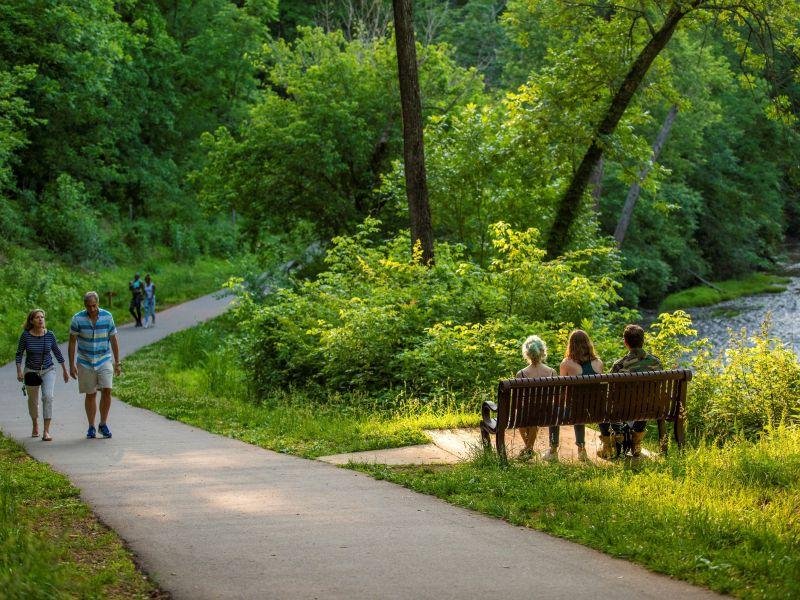 People walking on a trail alongside a creek, with three people sitting on a bench nearby, surrounded by lush green trees and vegetation in a park.
