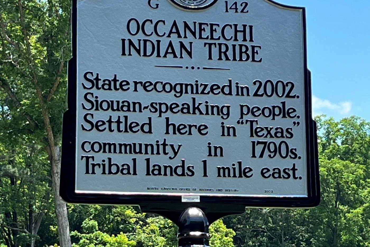 A historical marker sign for the Oceanechi Indian Tribe, recognizing the tribe in Texas, established in 2002. The sign notes the tribe's Siouan language, settlement in the 1790s, and lands located 1 mile east, with a background of green trees and a blue sky.