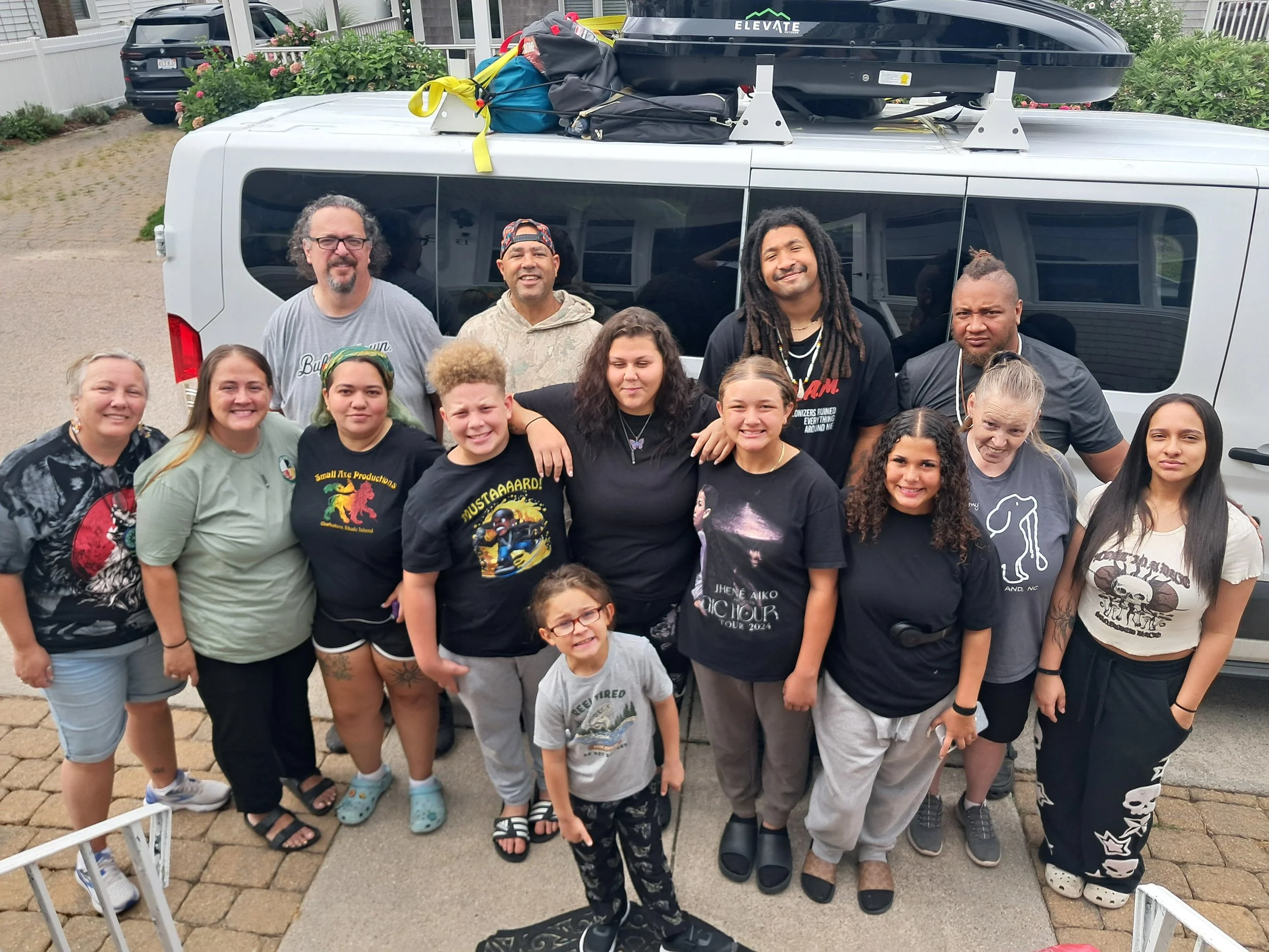 A group of 17 people, of various ages, standing together outdoors in front of a white van with a rooftop cargo box and camping gear, smiling at the camera.