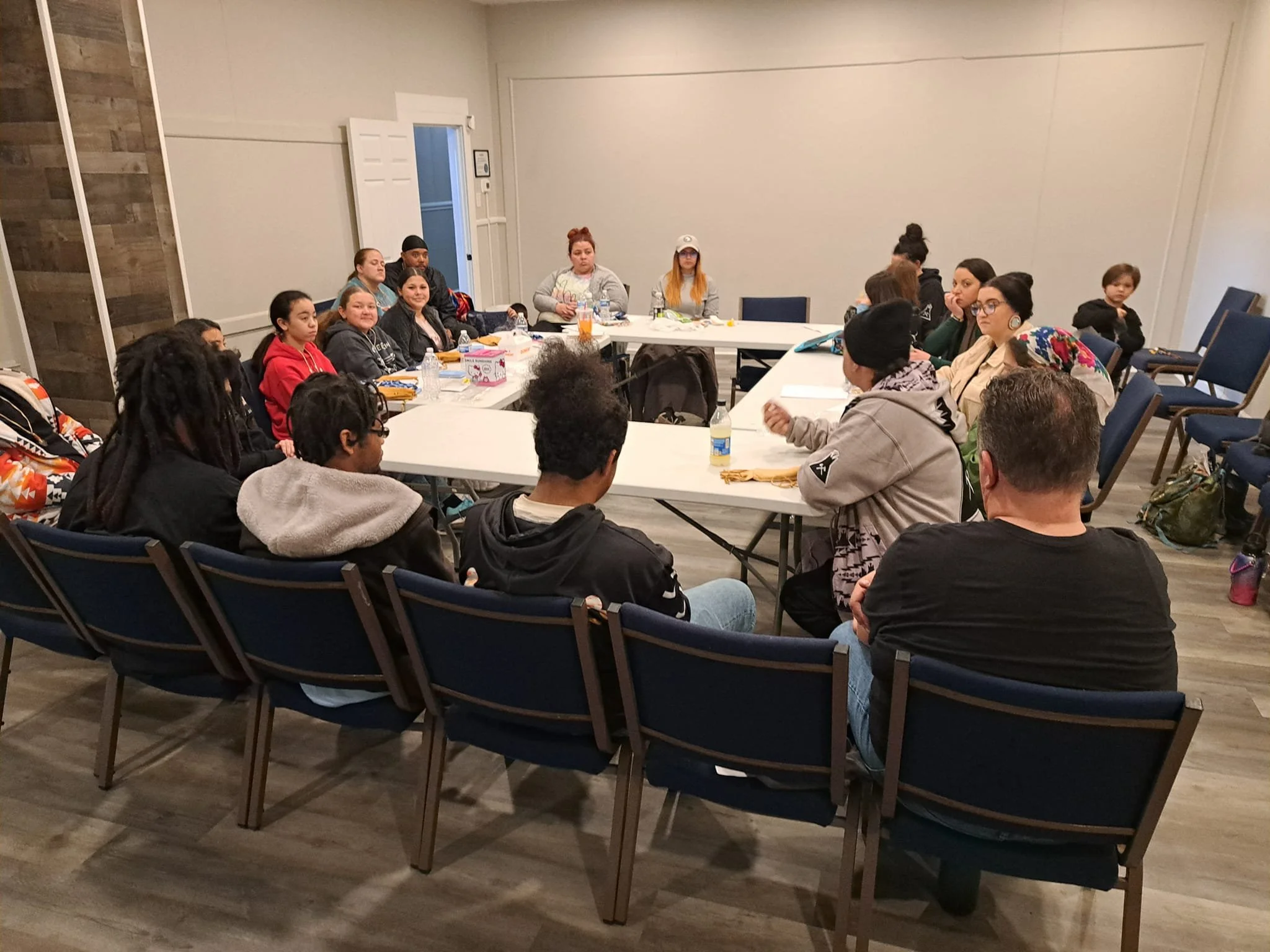 People gathered around a U-shaped conference table, listening attentively in a room with a beige wall and wooden paneling.