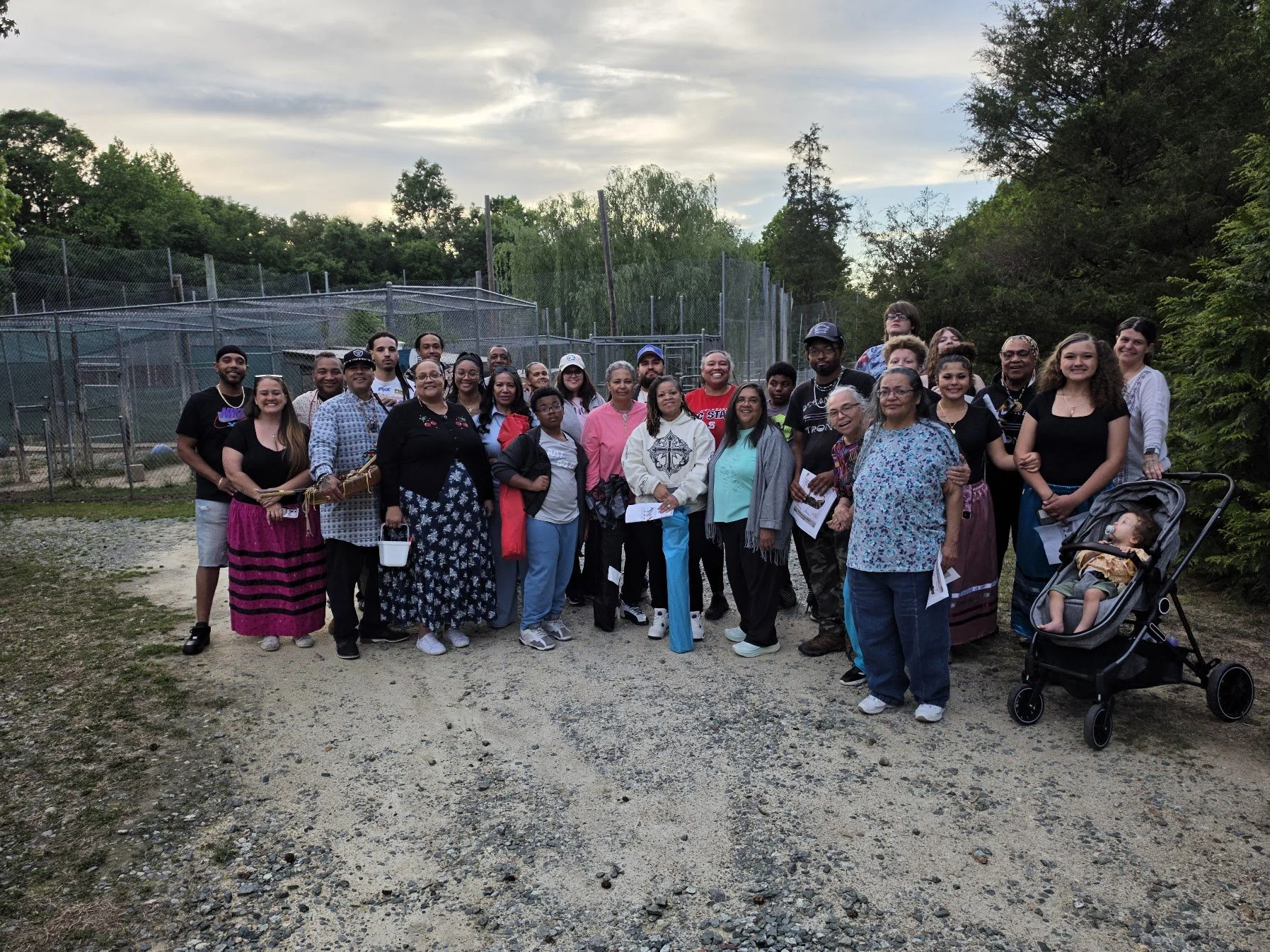 Group of diverse people standing outdoors for a photo, some holding papers or bags, with trees and a chain-link fence in the background.