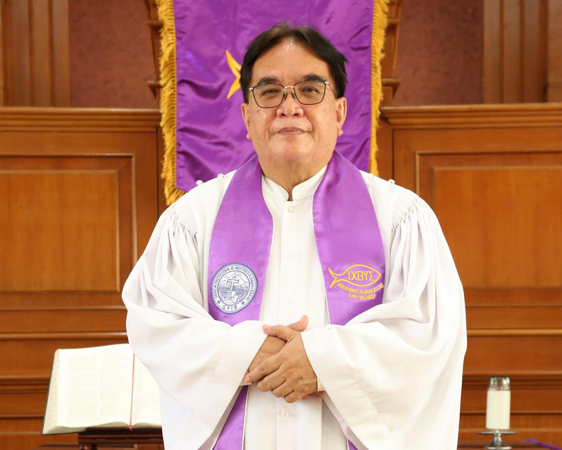 A man in a white religious robe with a purple stole, standing in front of a wooden background with a purple banner. The man is smiling and has his hands clasped in front of him.